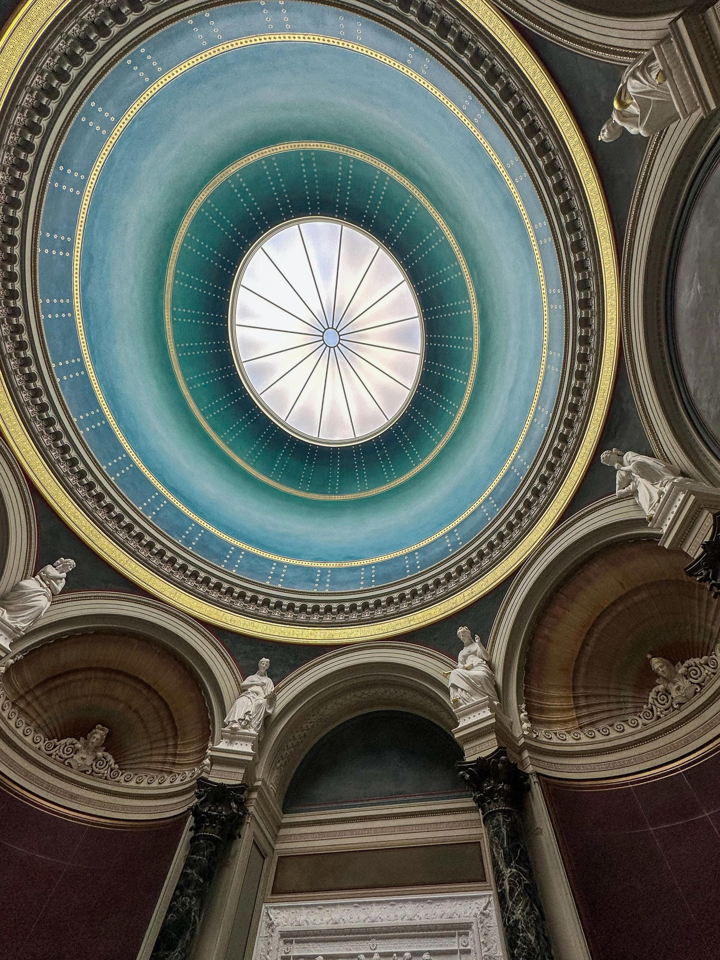 Looking up at a domed ceiling with a circular skylight at the center, surrounded by ornate architectural details and statues.