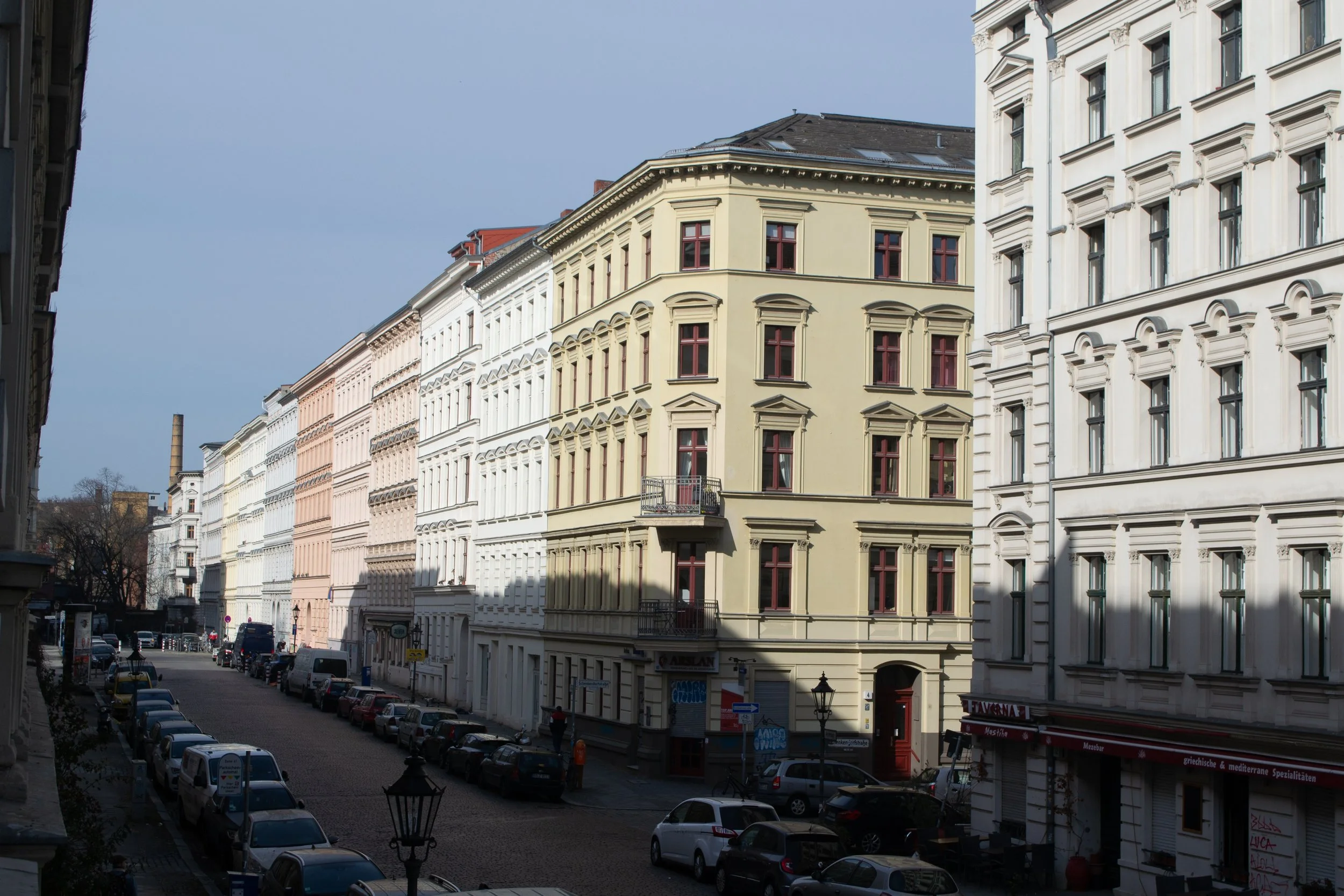 City street view with multi-story buildings, parked cars, a cloudy sky, and shadows cast on the buildings.