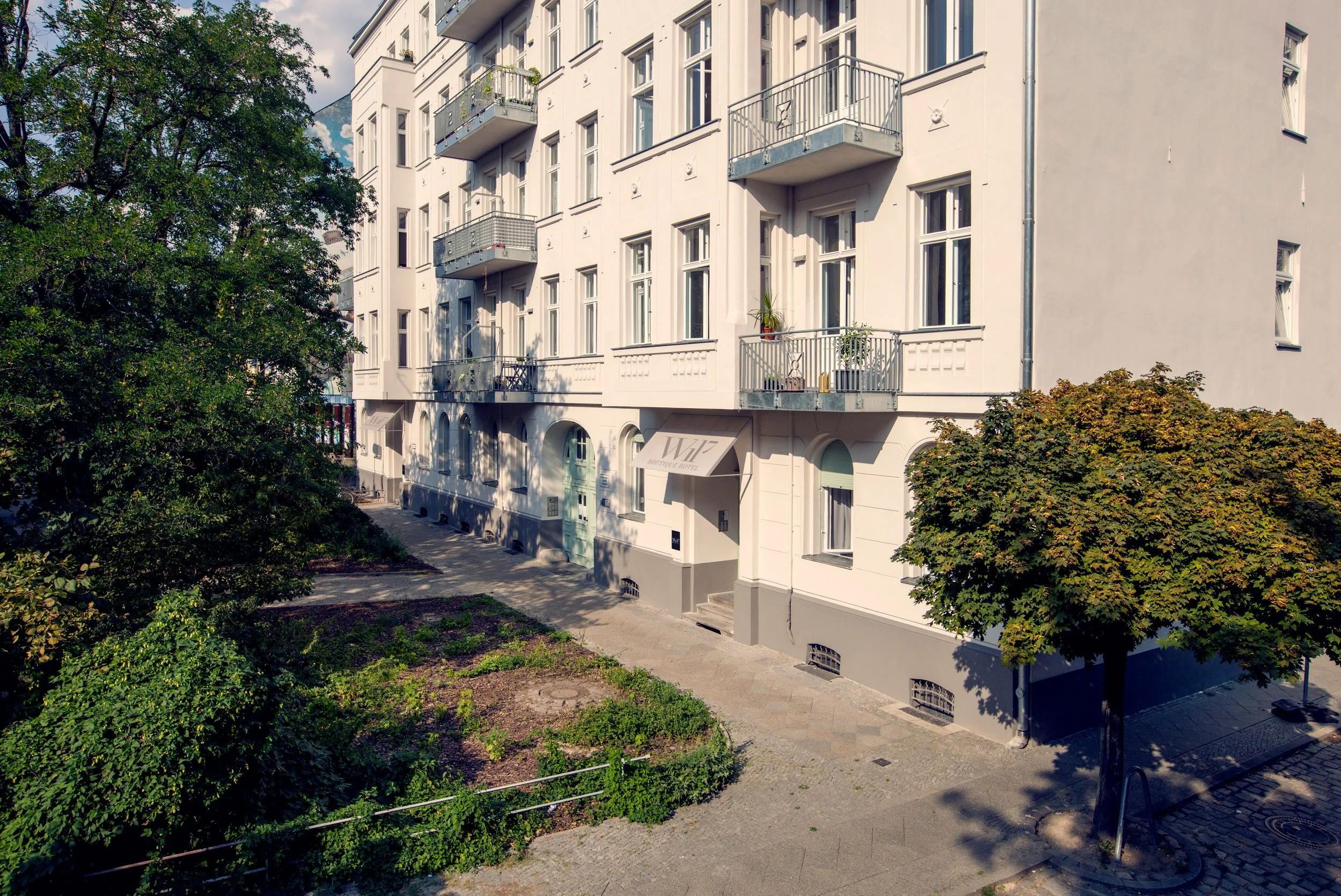 White multi-story residential building with balconies, potted plants on balconies, and a sign that reads "Whaz Boutique Hotel" above the entrance. There are trees and a small garden area in front, with a pathway and stairs leading to the entrance.