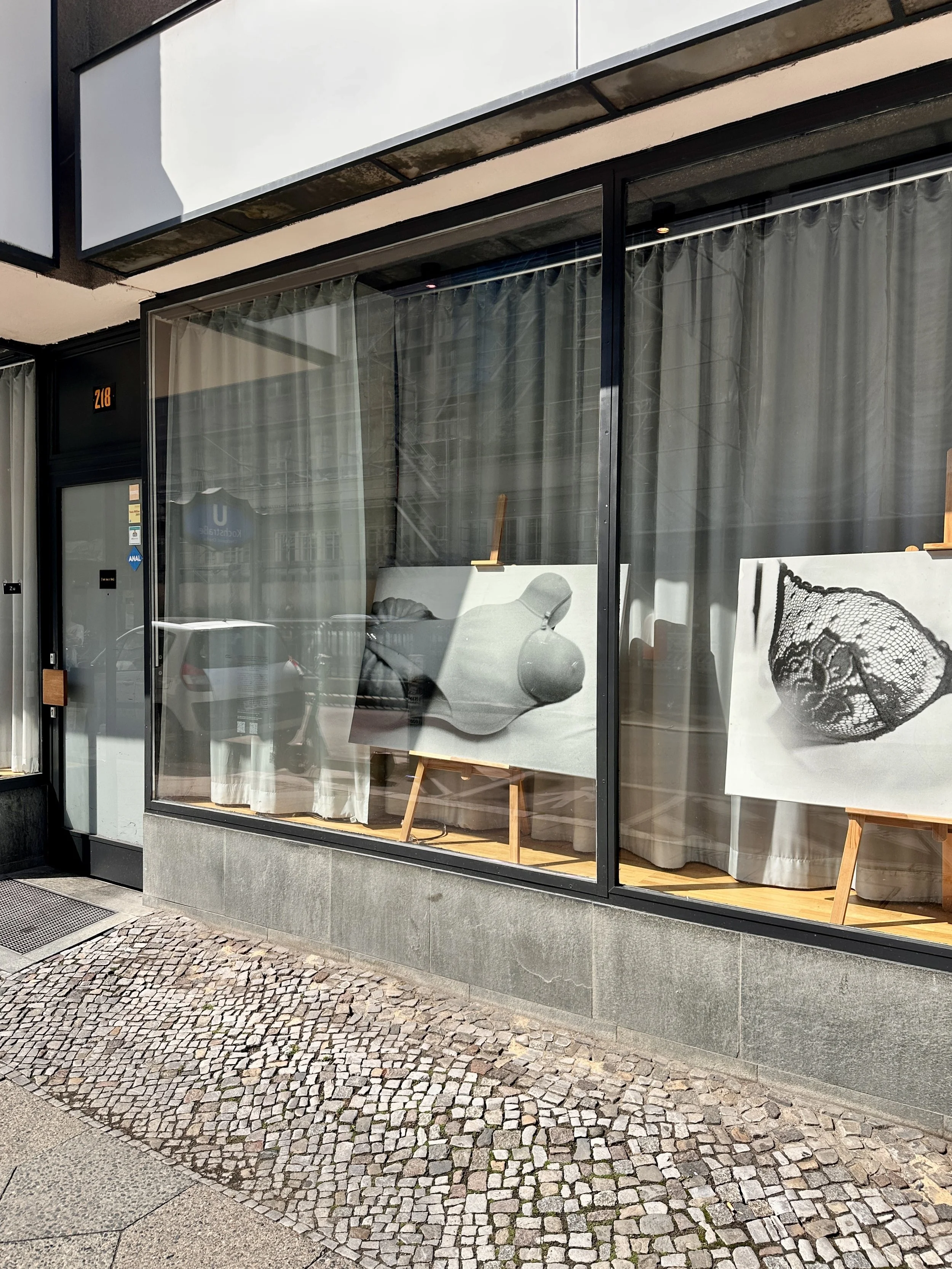 Storefront window with artwork of a black and white photo of a woman wearing a lace hat, displayed on an easel inside the shop. The shop has a modern exterior with black framing and a cobblestone sidewalk outside.