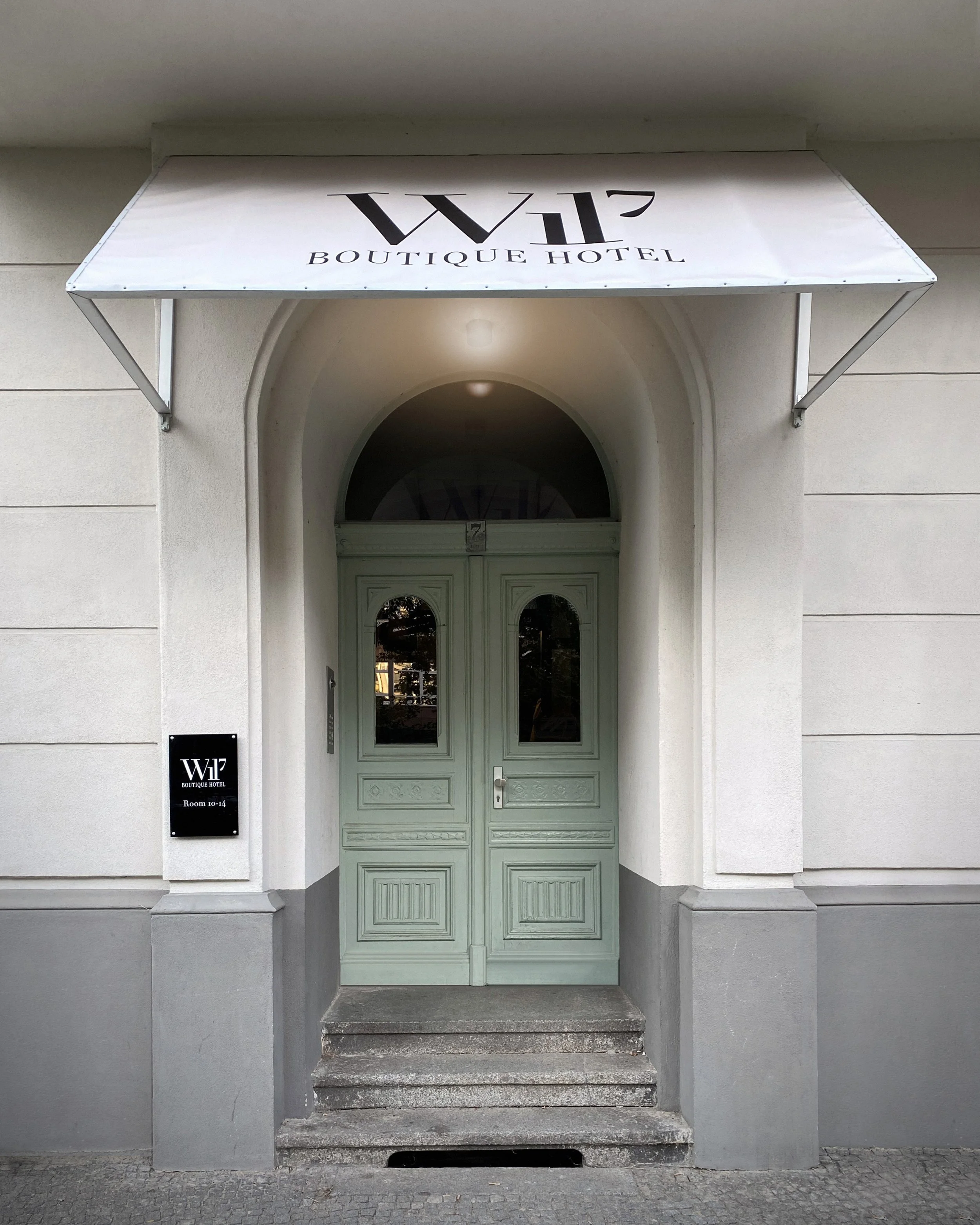 Green double doors with decorative panels and arched glass windows, entrance to W17 Boutique Hotel, with a black sign on the left indicating rooms 10-14, and a white awning overhead with the hotel logo.