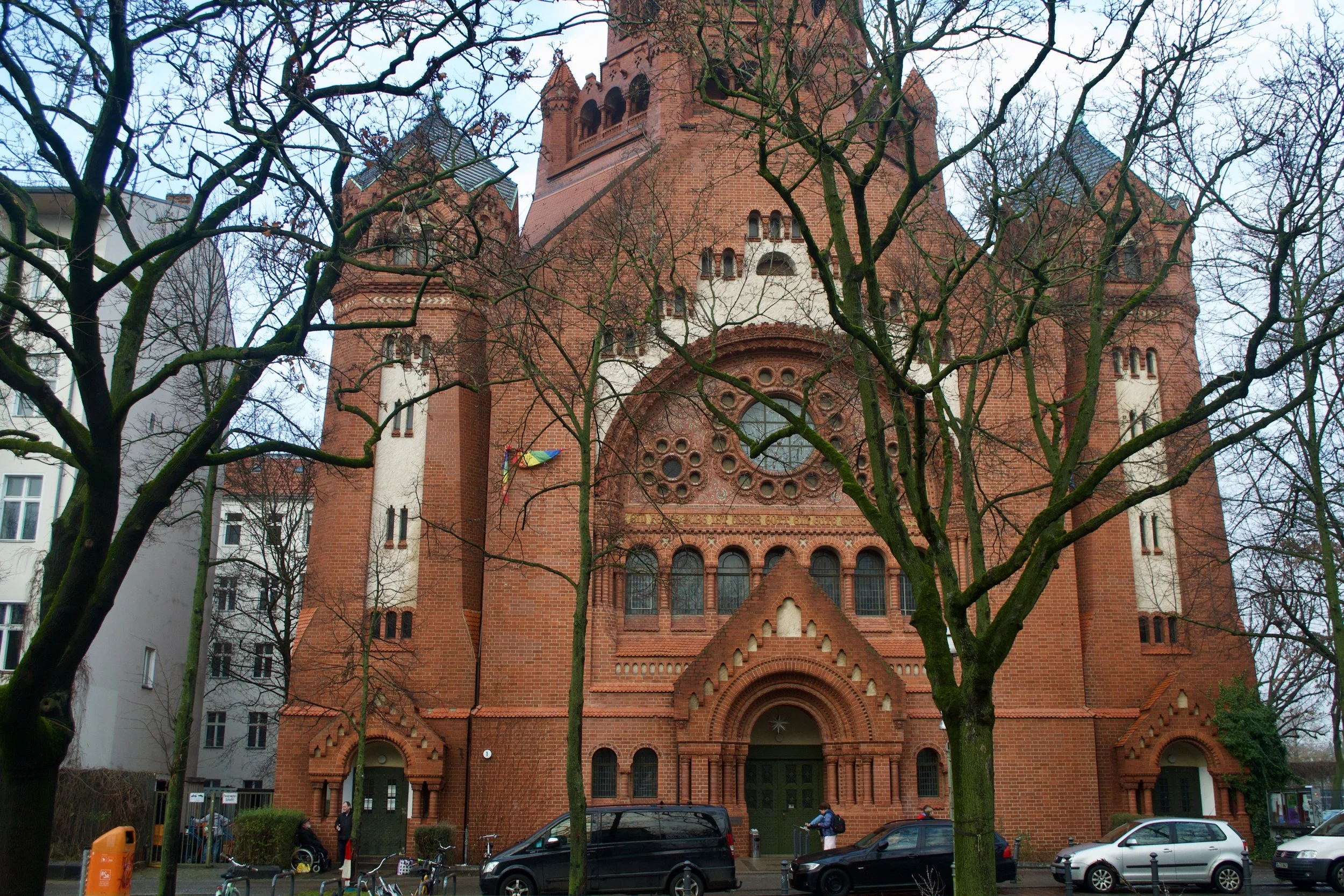 A large red brick church with gothic architectural features, tall towers, arched windows, and decorative circular patterns on the facade, seen through leafless trees on a rainy day, with cars and people outside.