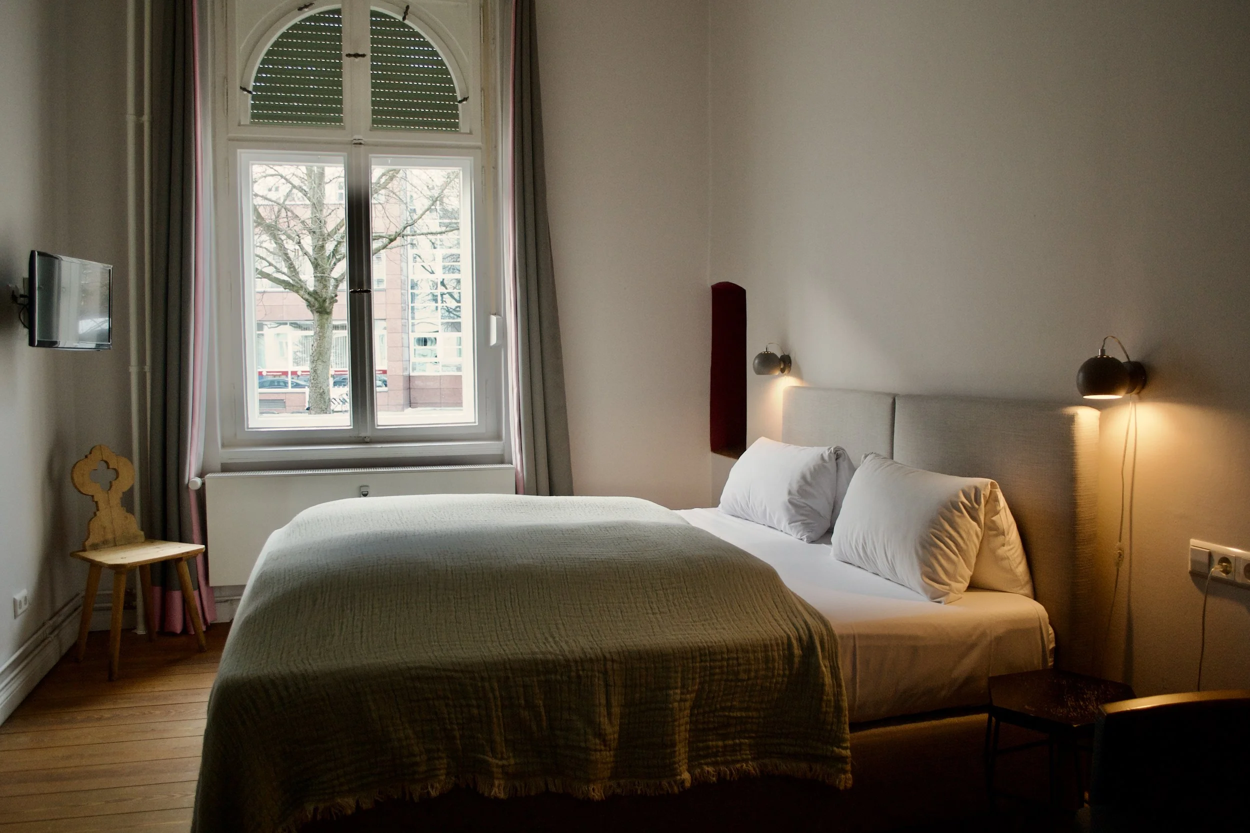 A bedroom with a large window showing a tree outside, beige walls, a bed with a beige headboard and white bedding, two bedside lamps, a small wooden chair, and a flat-screen TV mounted on the wall.