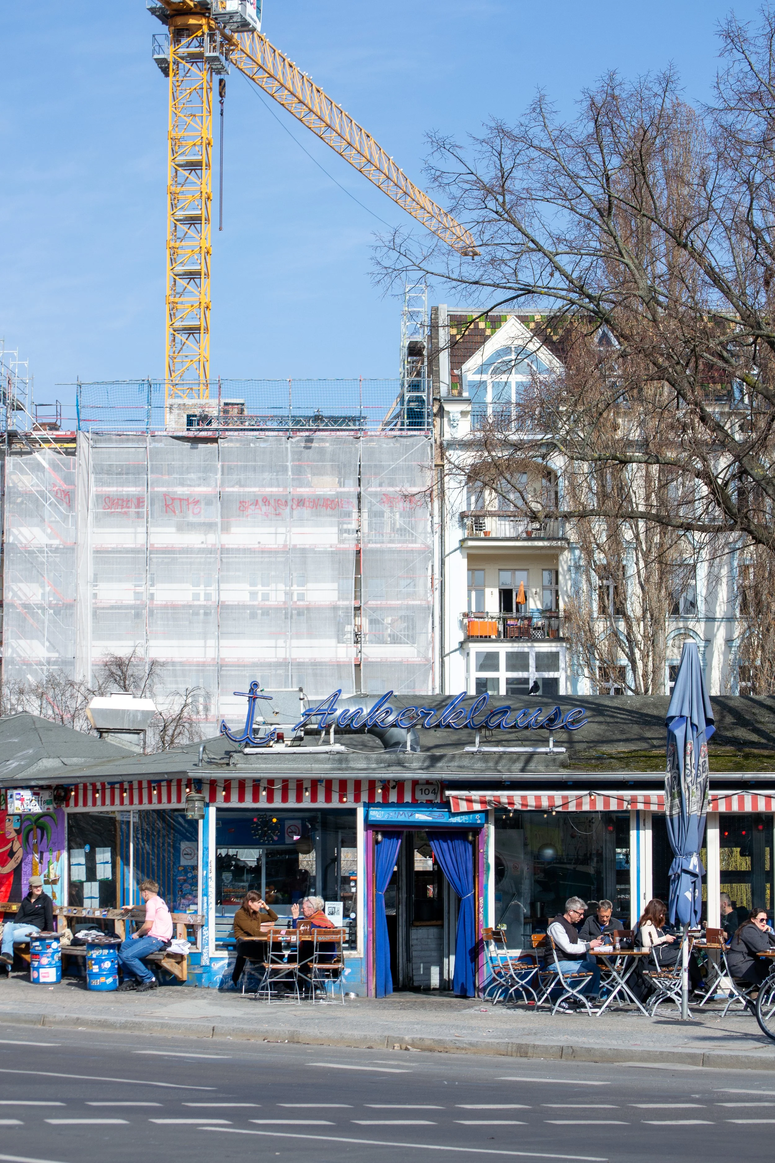 A cafe named 't Ankerklause with outdoor seating, people dining, and a construction site with buildings and a yellow crane behind it.