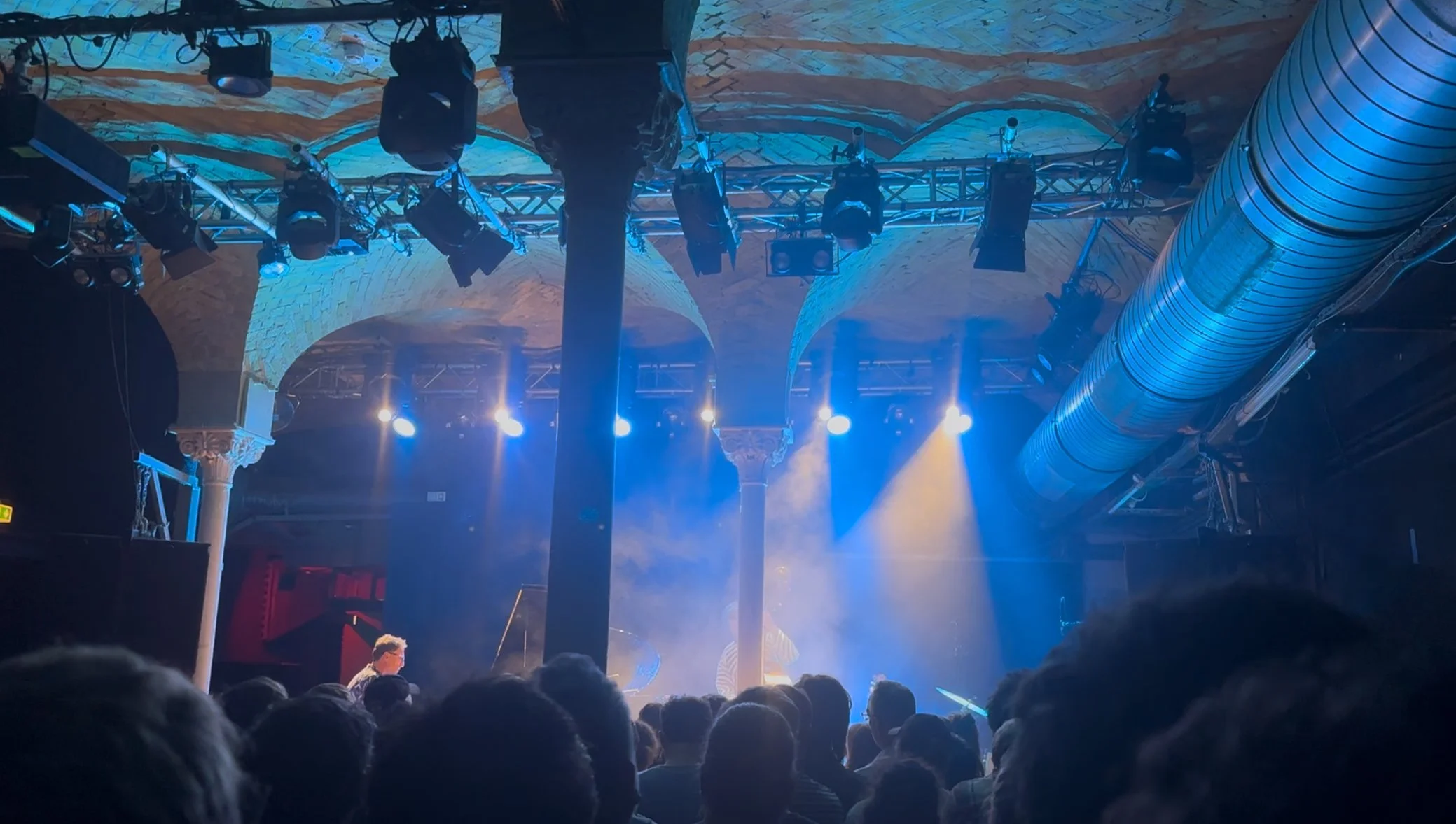 A concert or performance on a stage with blue and yellow lighting, an audience in front, and stage equipment including lights and ventilation ducts overhead.
