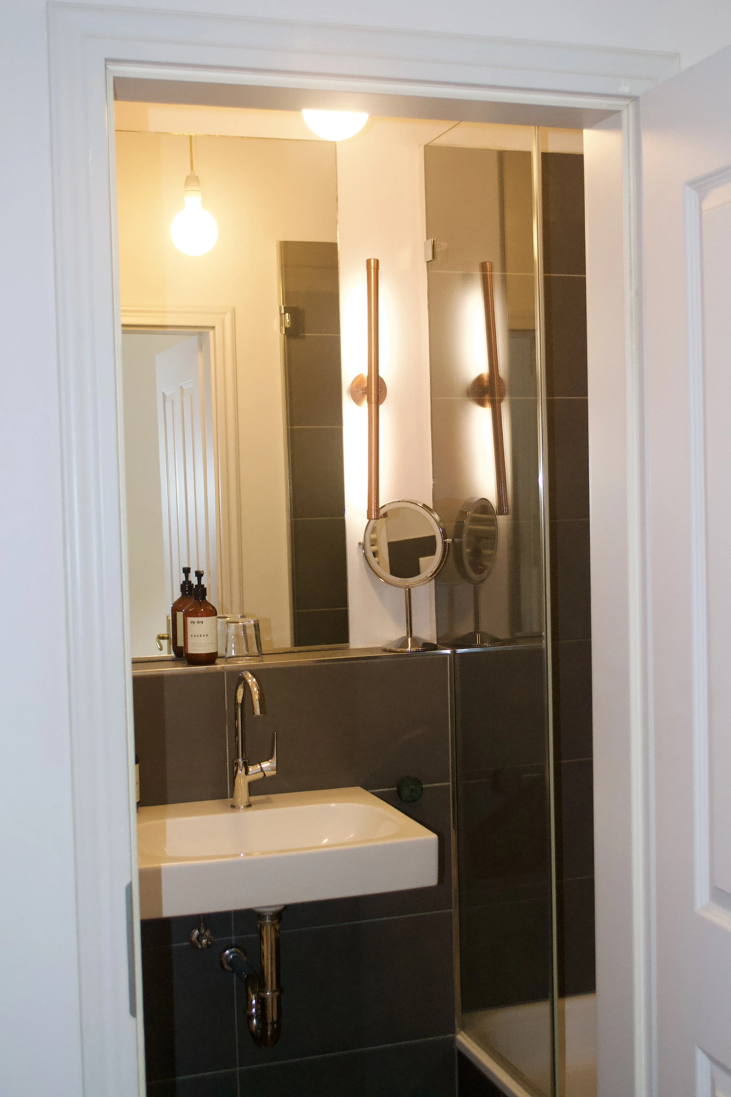 A bathroom seen through a doorway with a white frame. Inside, there is a mirror above a sink, with copper-colored wall sconce lights reflecting in the mirror. There are bottles of soap on the sink counter, and a faucet. To the right, there is a showe