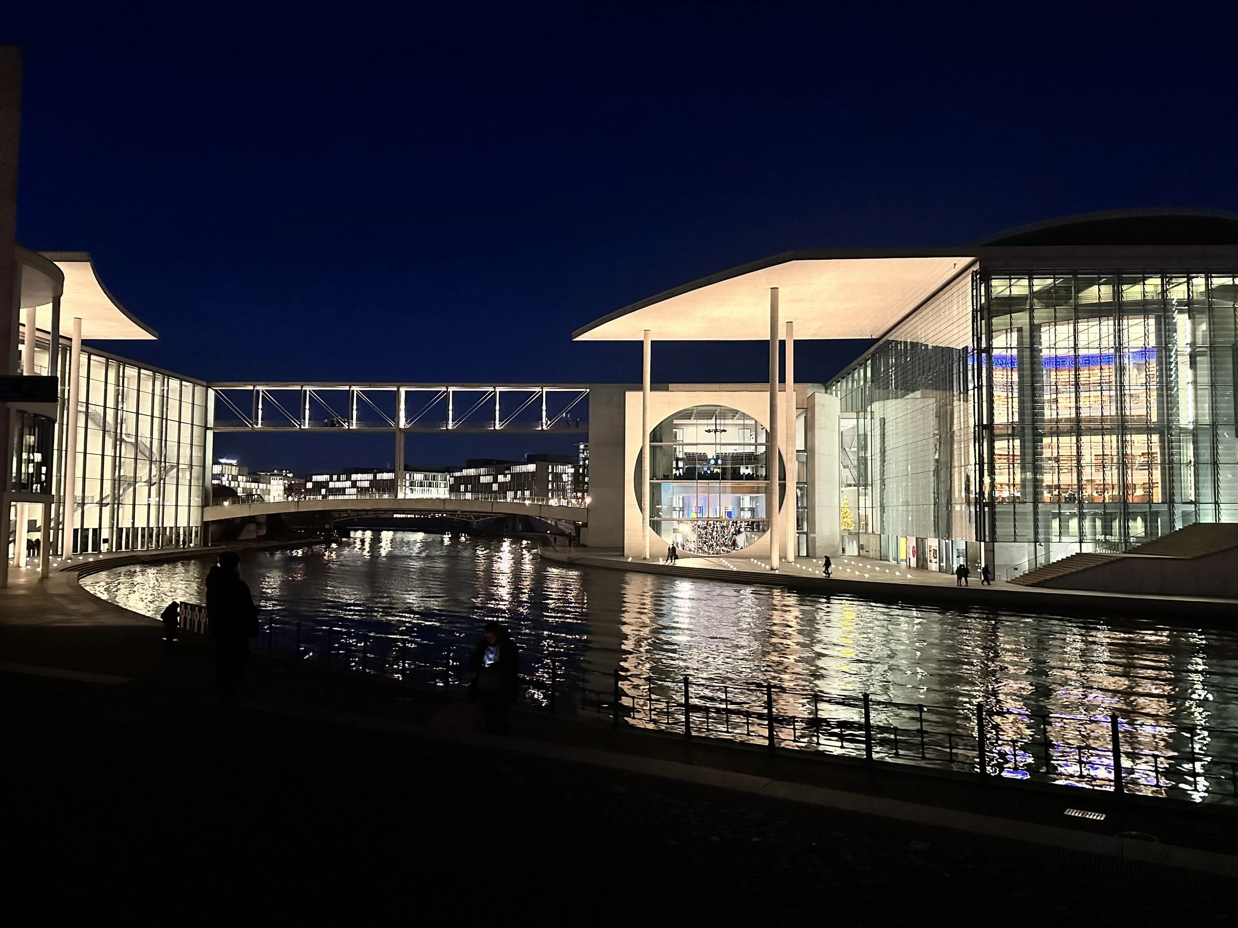 Night view of a modern architectural building with glass walls and reflections on the water in front, with a dark blue sky and several silhouetted people walking along the water.