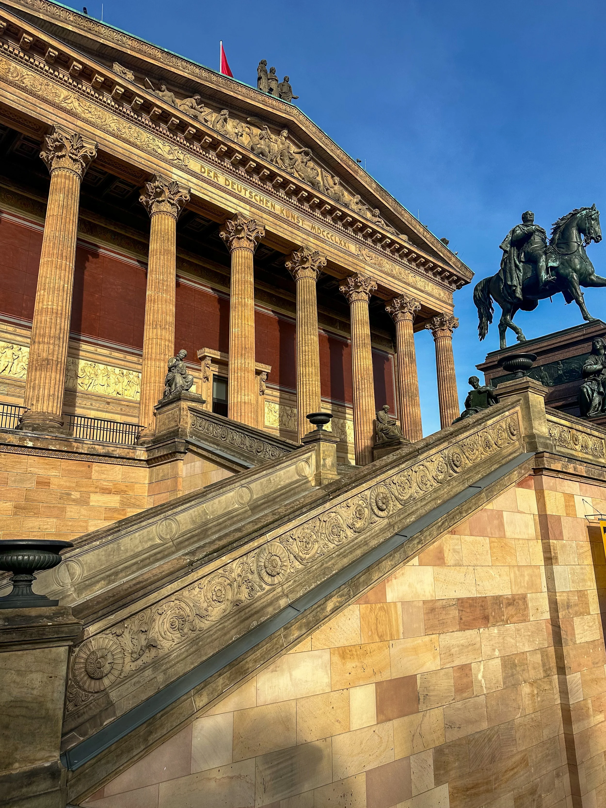 Ancient Greek-style building with tall columns, a staircase with ornate carvings, statues, and a large equestrian statue, against a clear blue sky.