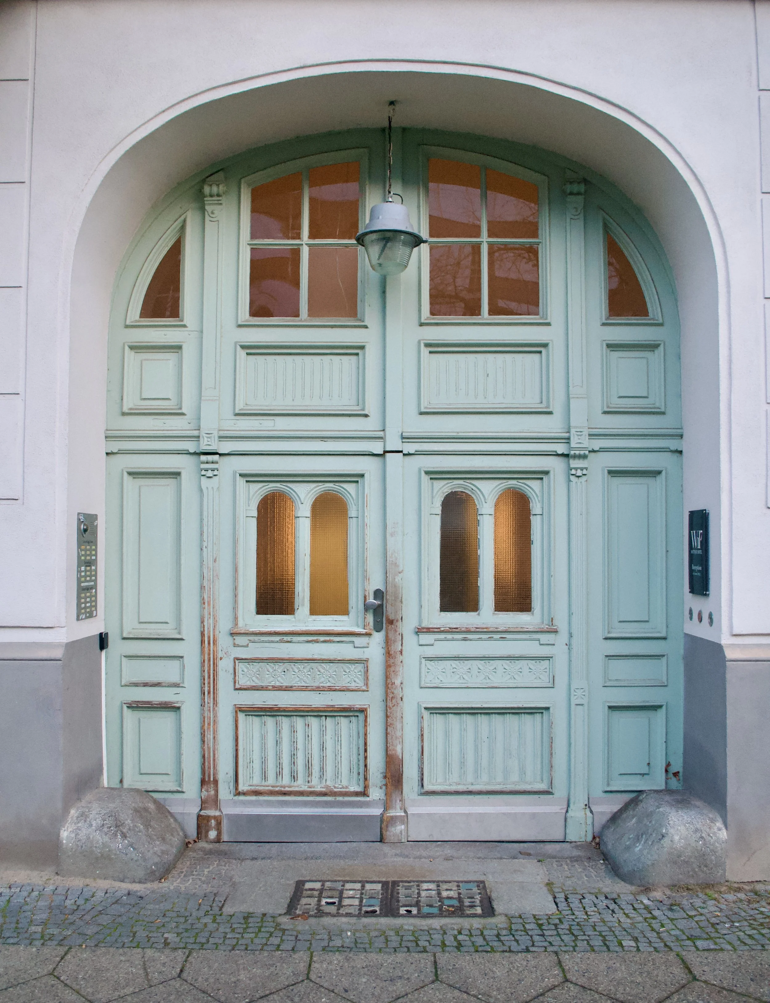 Old wooden double door painted in faded light green, with arched windows and decorative panels, set in a building with an arched entrance and exterior lantern, flanked by concrete barriers.