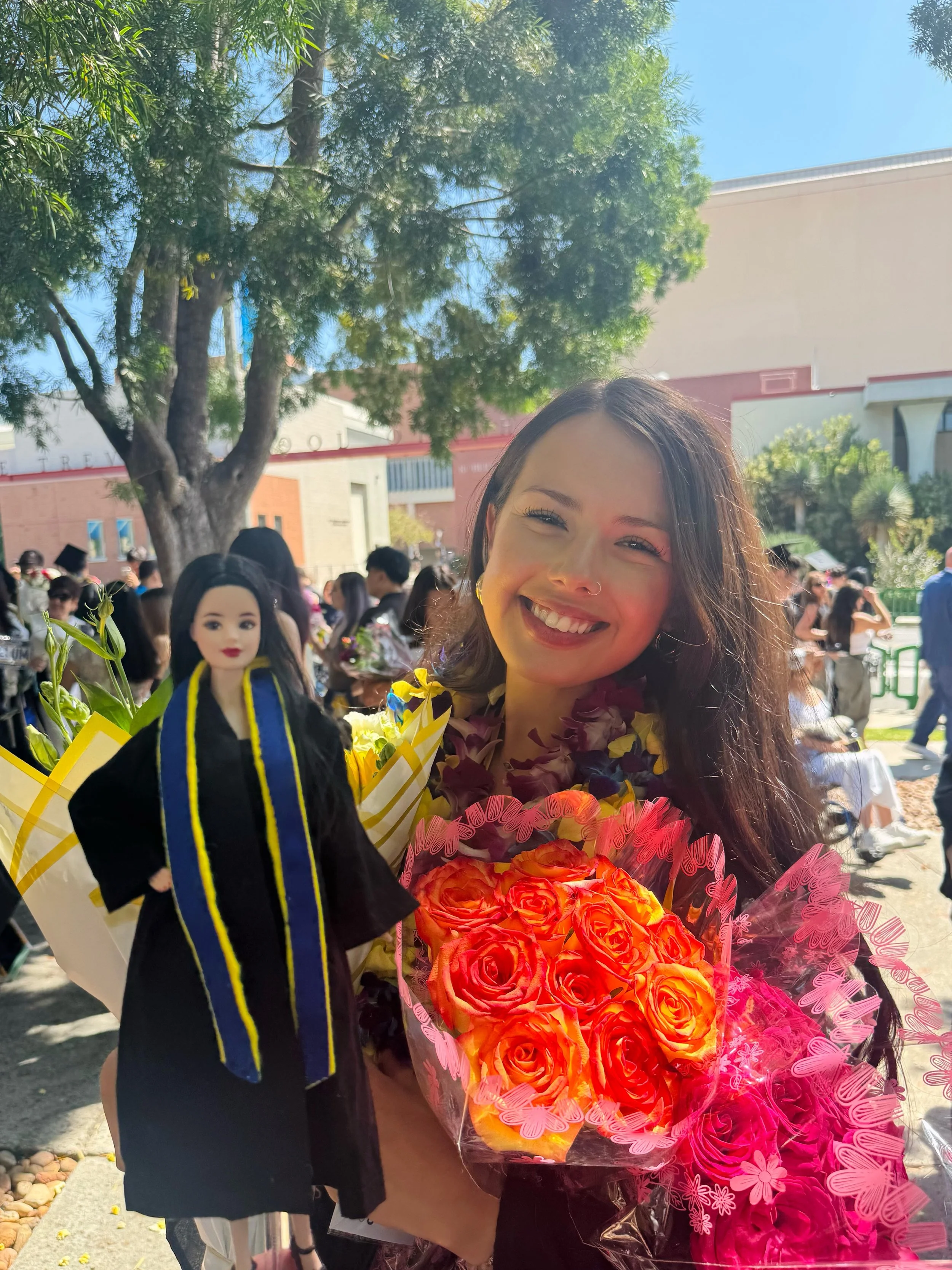 A woman with long brown hair and a big smile, wearing a flower lei, holding a bouquet of orange and pink roses, standing among a crowd at an outdoor graduation celebration.