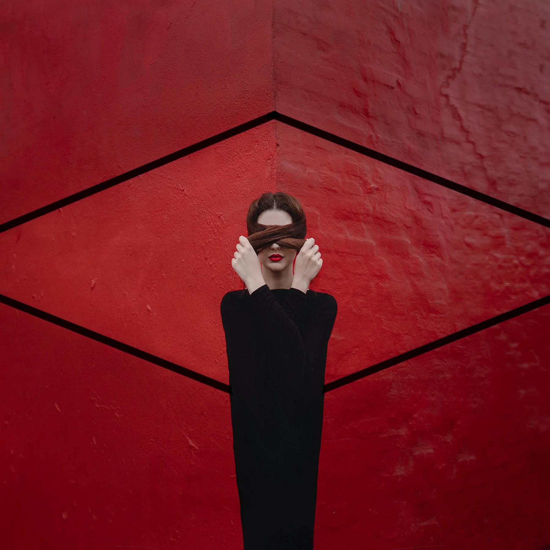 A woman in a black long dress with red lips stands in the center against a red wall with black lines forming a diamond. She covers her eyes with strands of long dark hair.