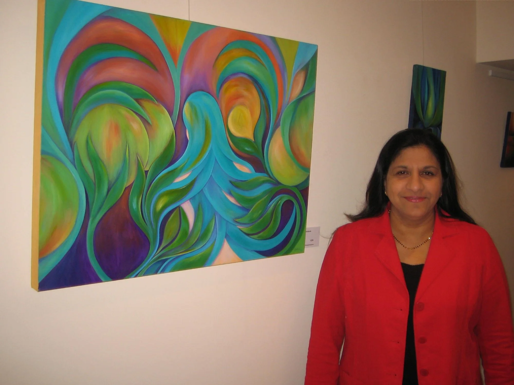 A woman in a red blazer standing in front of an abstract, colorful painting with swirling green, blue, purple, and orange patterns.