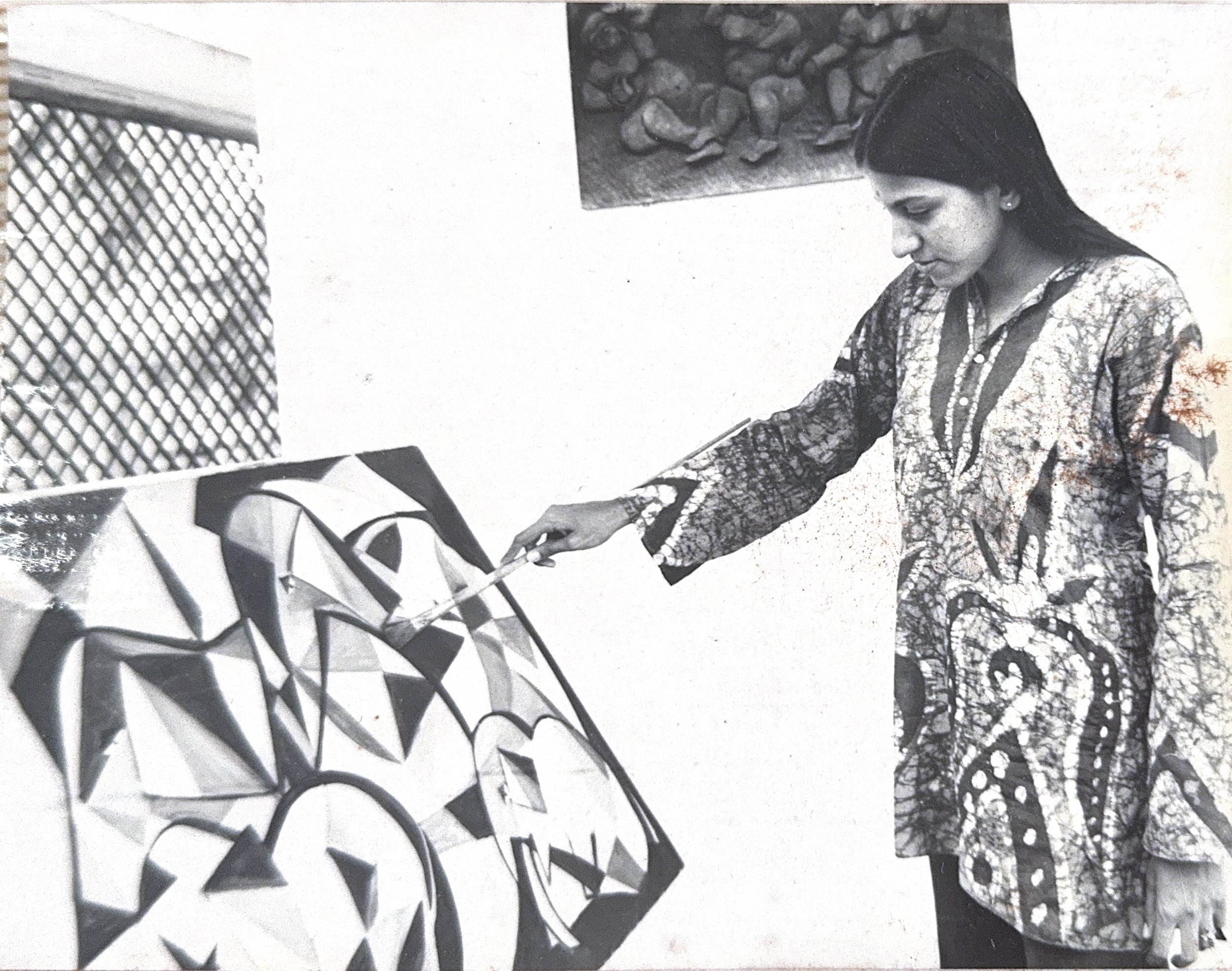 A woman wearing a patterned dress is painting an abstract geometric artwork on a canvas. She is holding a paintbrush and appears focused on her work.
