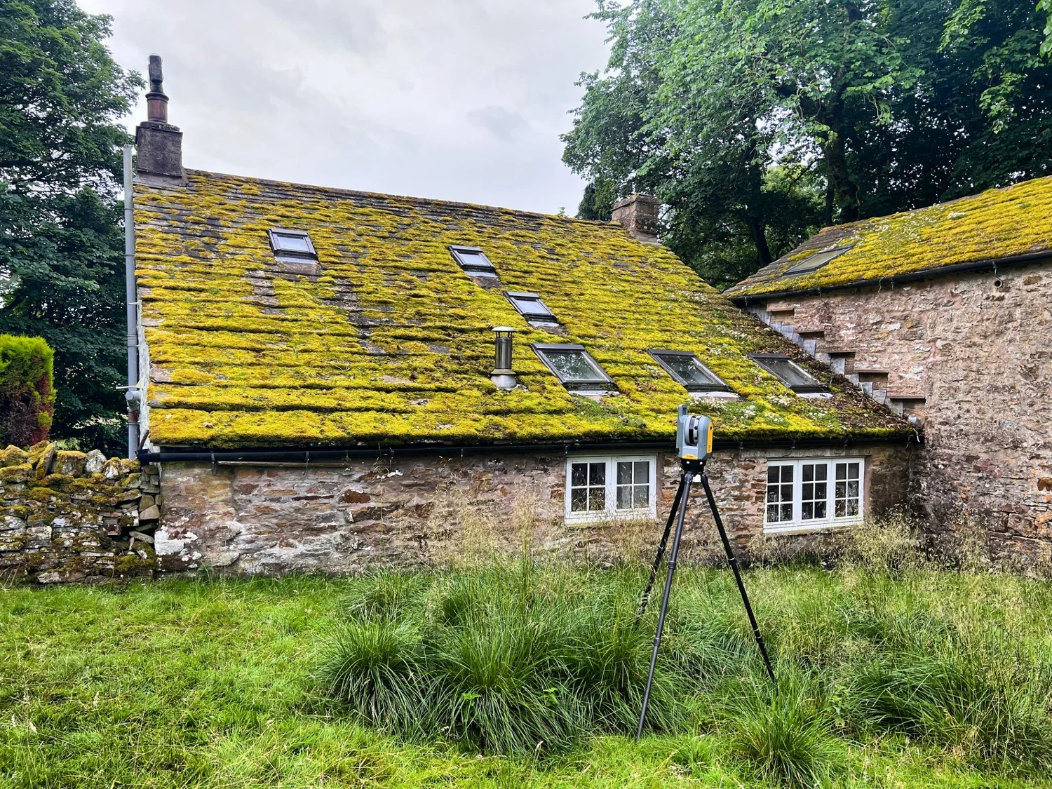 Laser scanner on a tripod in front of a moss-covered stone roof with multiple skylights
