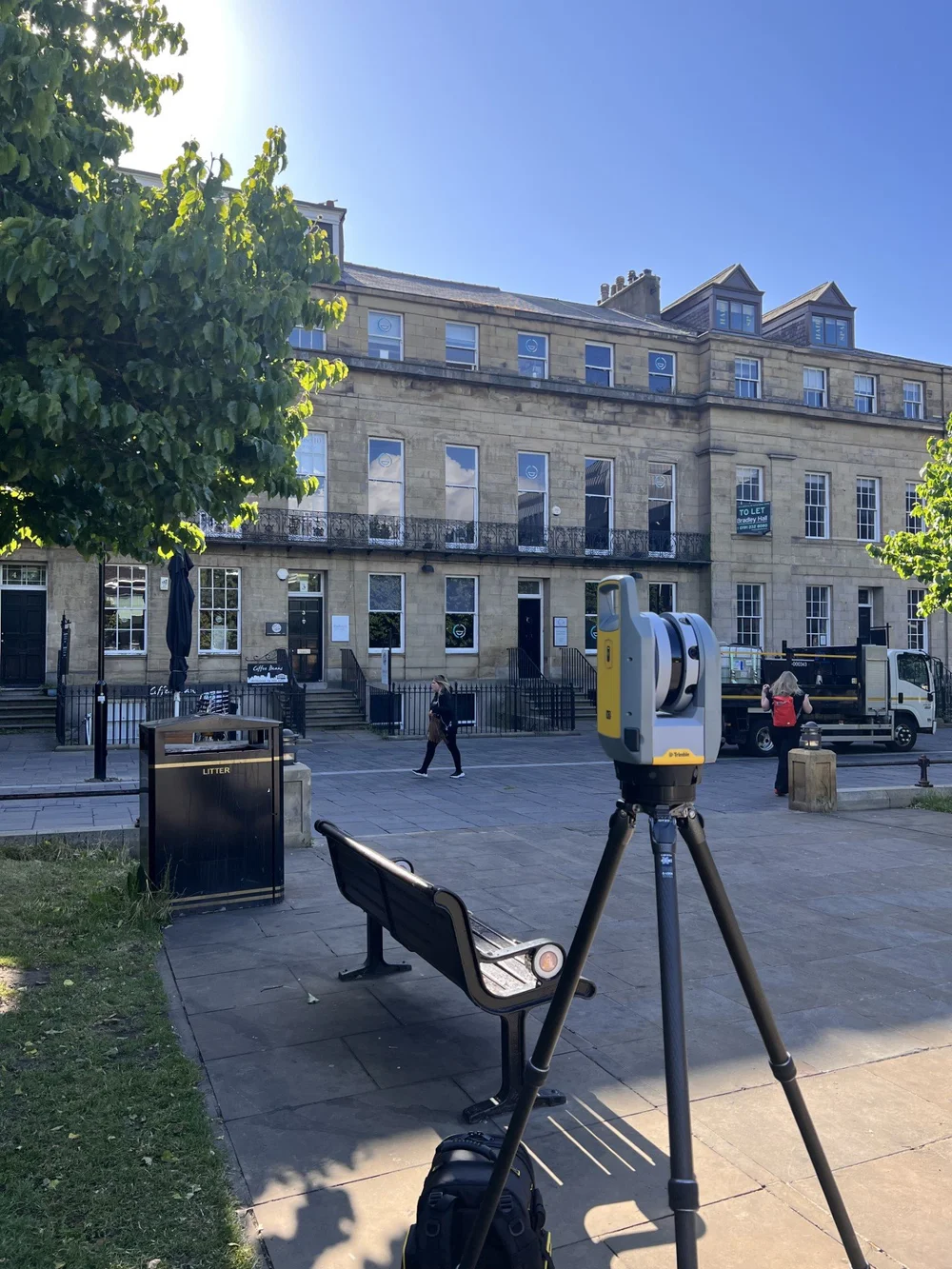 Tripod-mounted scanner on a street in front of a multi-storey stone building with shopfronts
