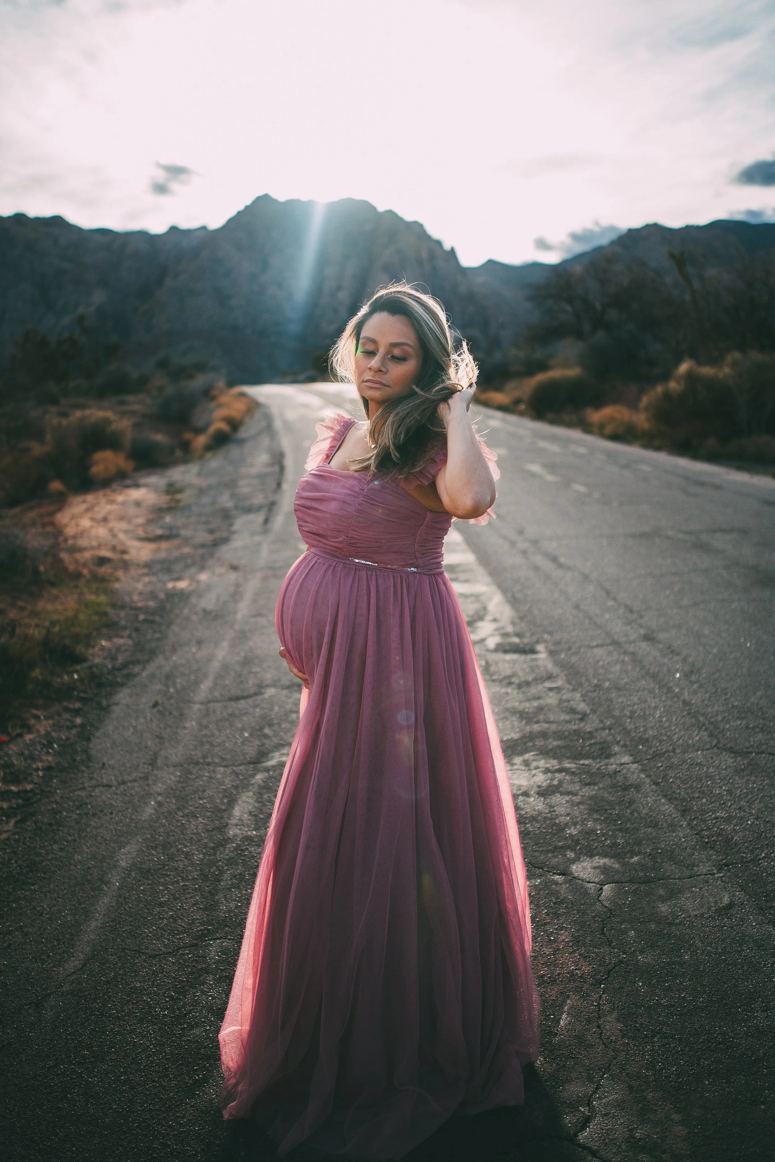Pregnant woman in a purple dress standing on a deserted road in a mountainous area during sunset.