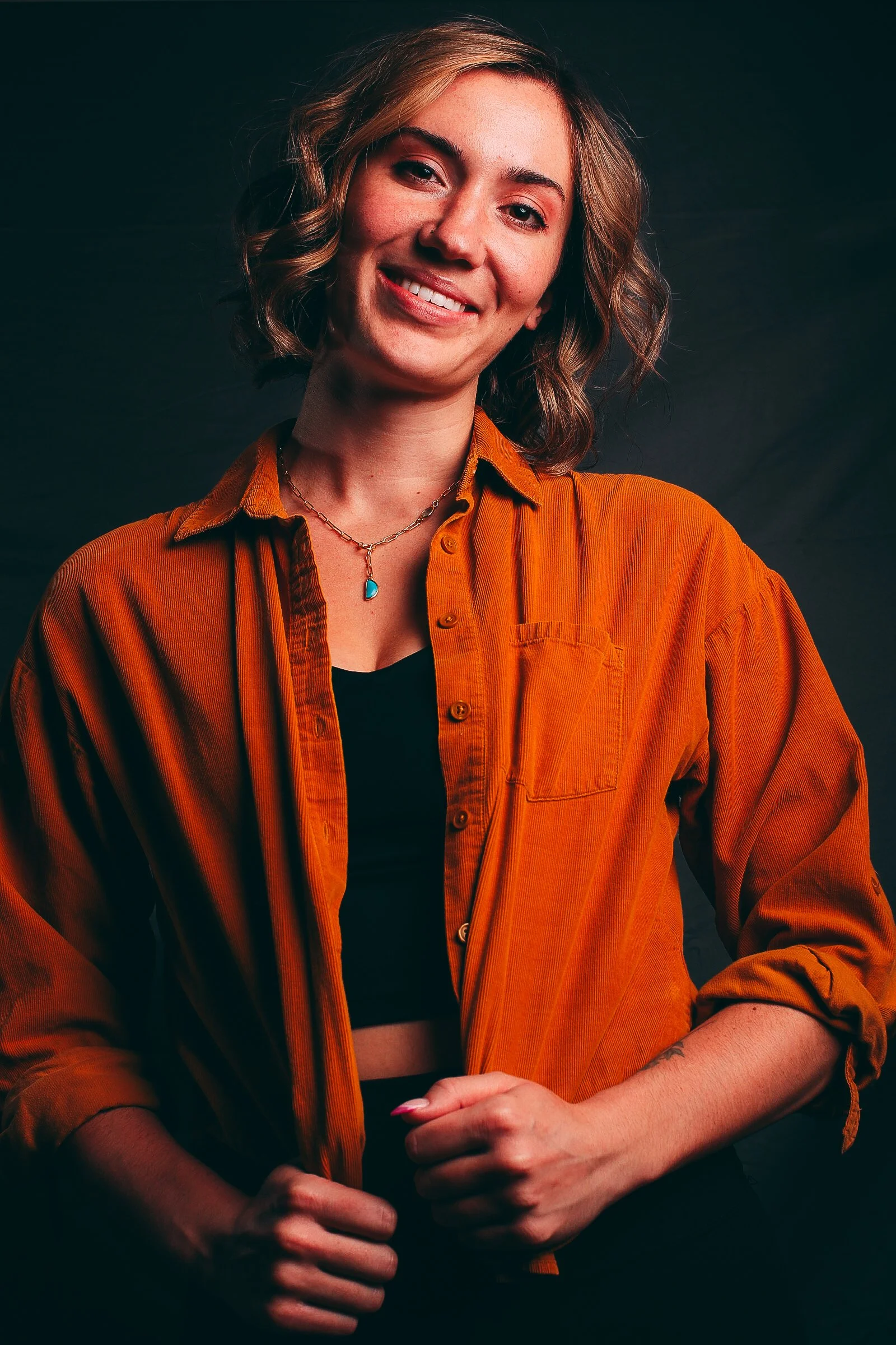 Portrait of a young woman with shoulder-length wavy hair smiling, wearing a black top, an orange shirt, a turquoise necklace, and nail polish, standing against a dark background.