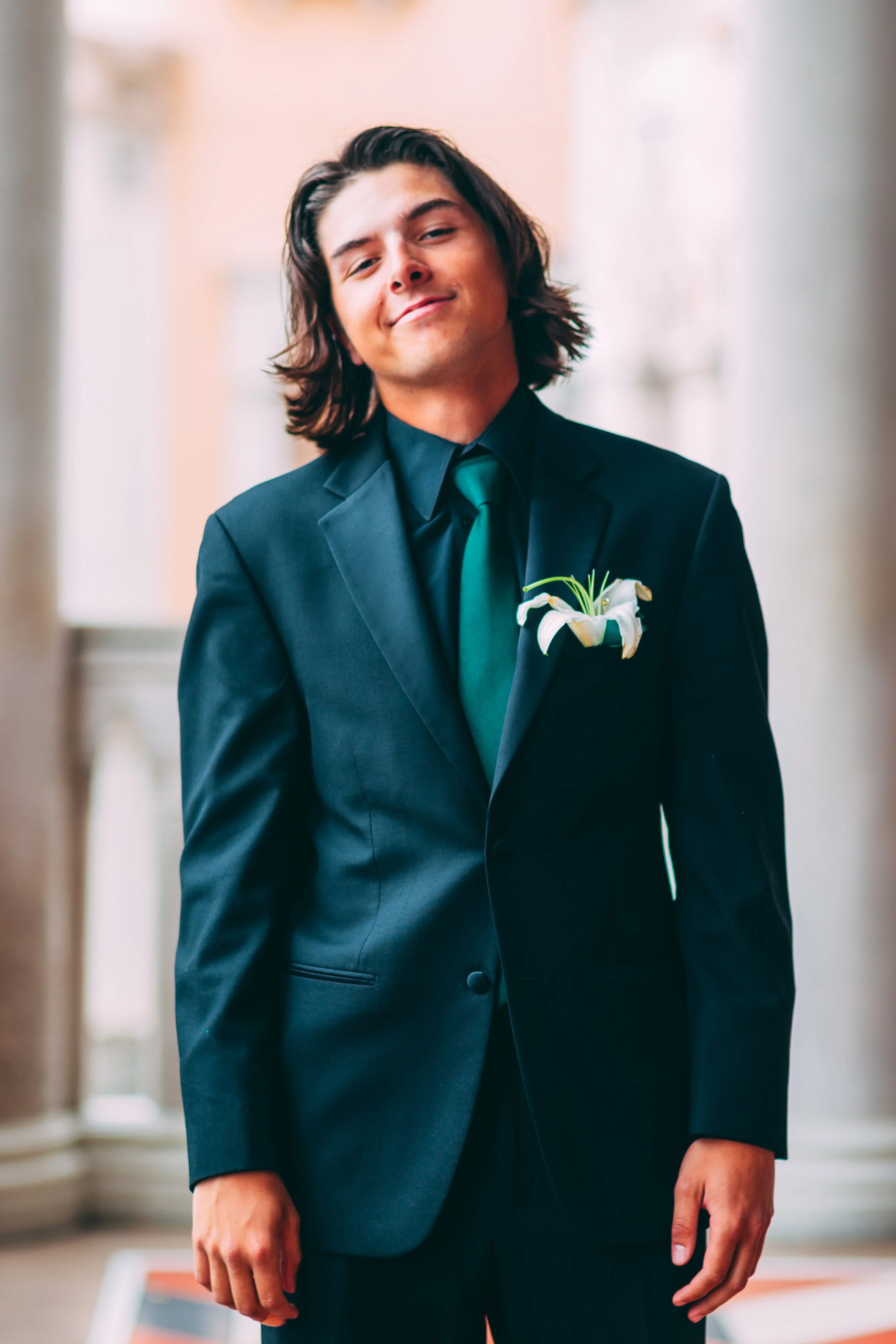 Young man smiling and dressed in a formal black suit and teal tie, with a white lily boutonniere on his lapel, standing outdoors.