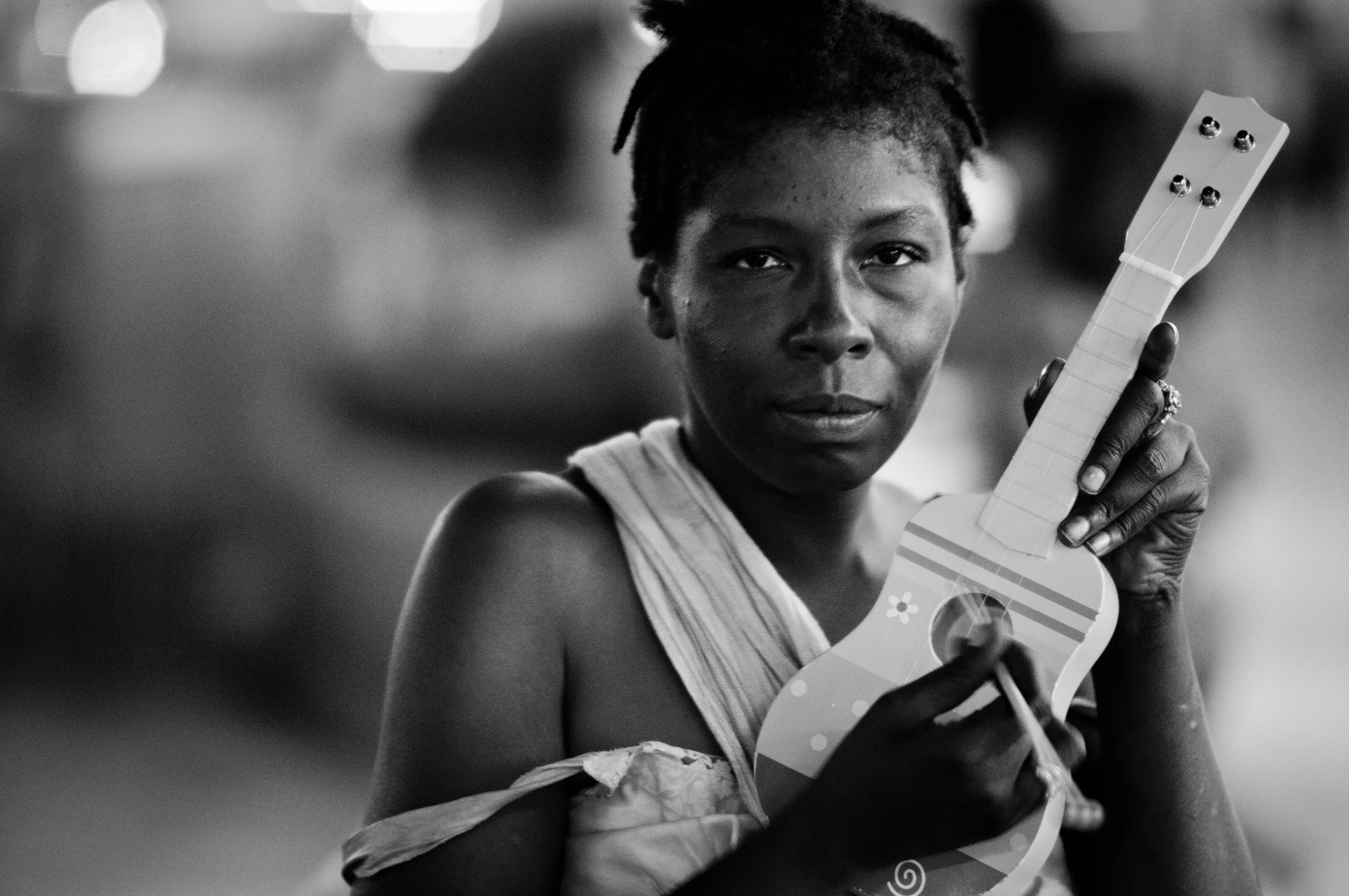 A young girl with short dreadlocks looking at the camera while holding a small, colorful toy guitar and a drumstick in her hand.