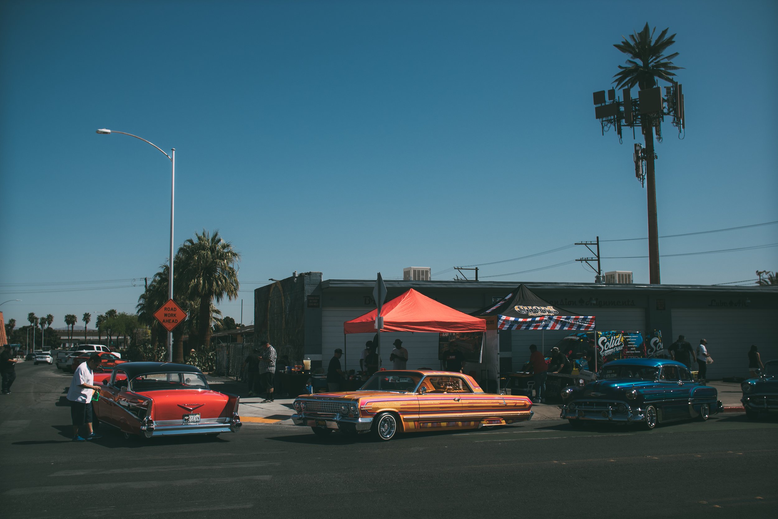 Vintage cars parked along a street during a car show with tents and people, palm trees, and a clear blue sky.