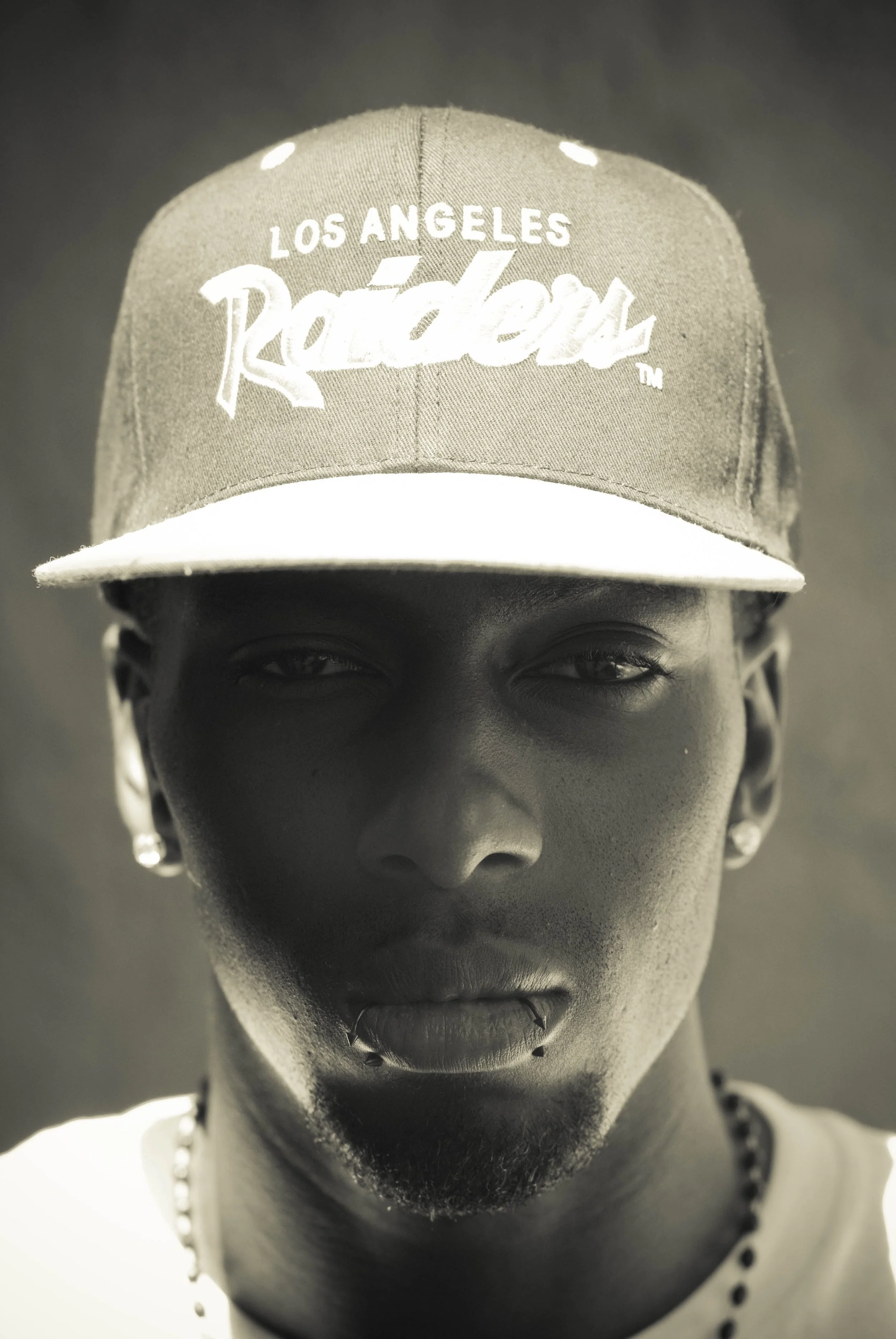 Close-up black and white portrait of a person wearing a Los Angeles Raiders baseball cap, with earrings, lips, and a necklace visible.