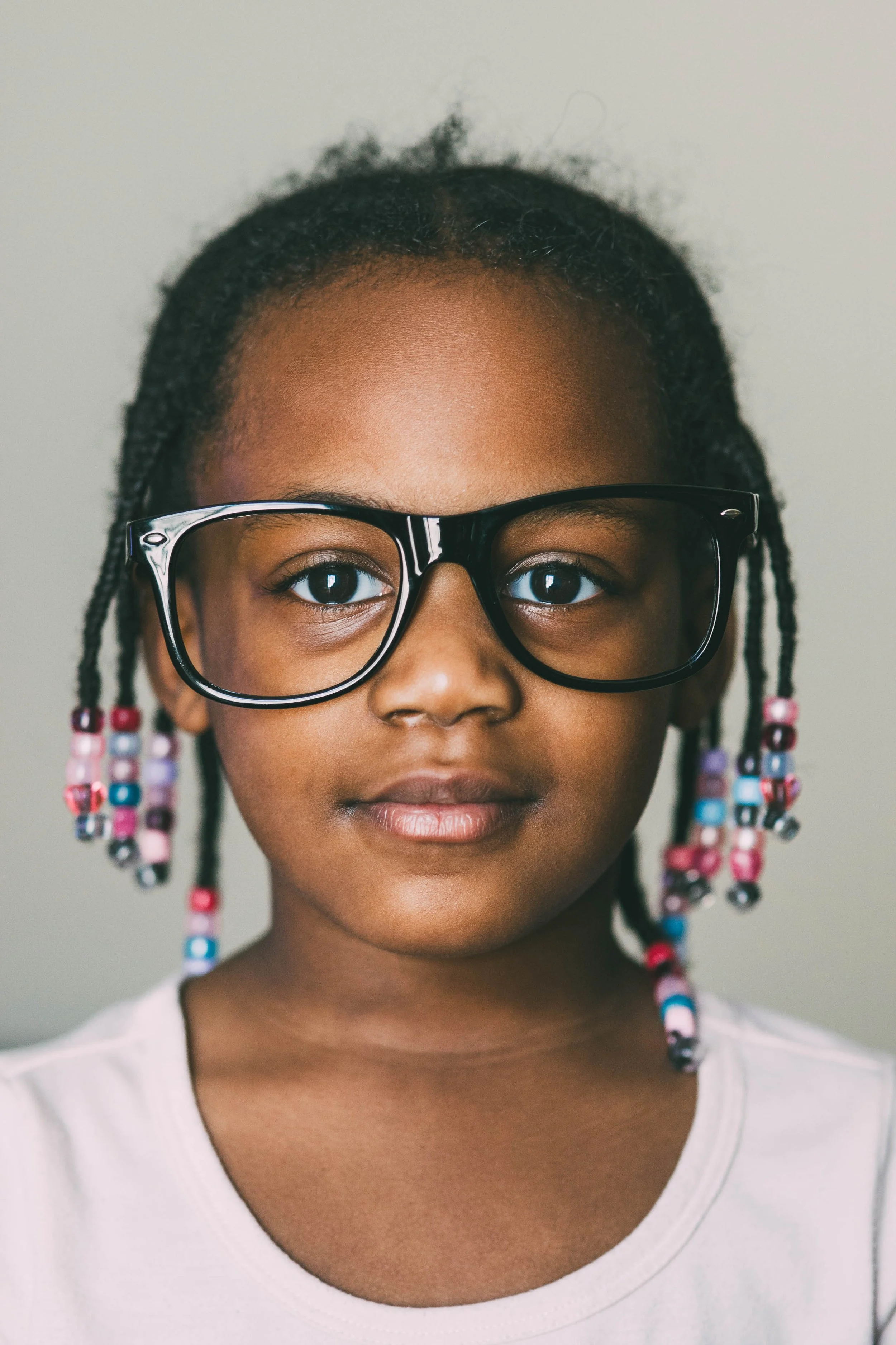 A young girl with dark skin and colorful beaded hair, wearing large black glasses, looking directly at the camera.