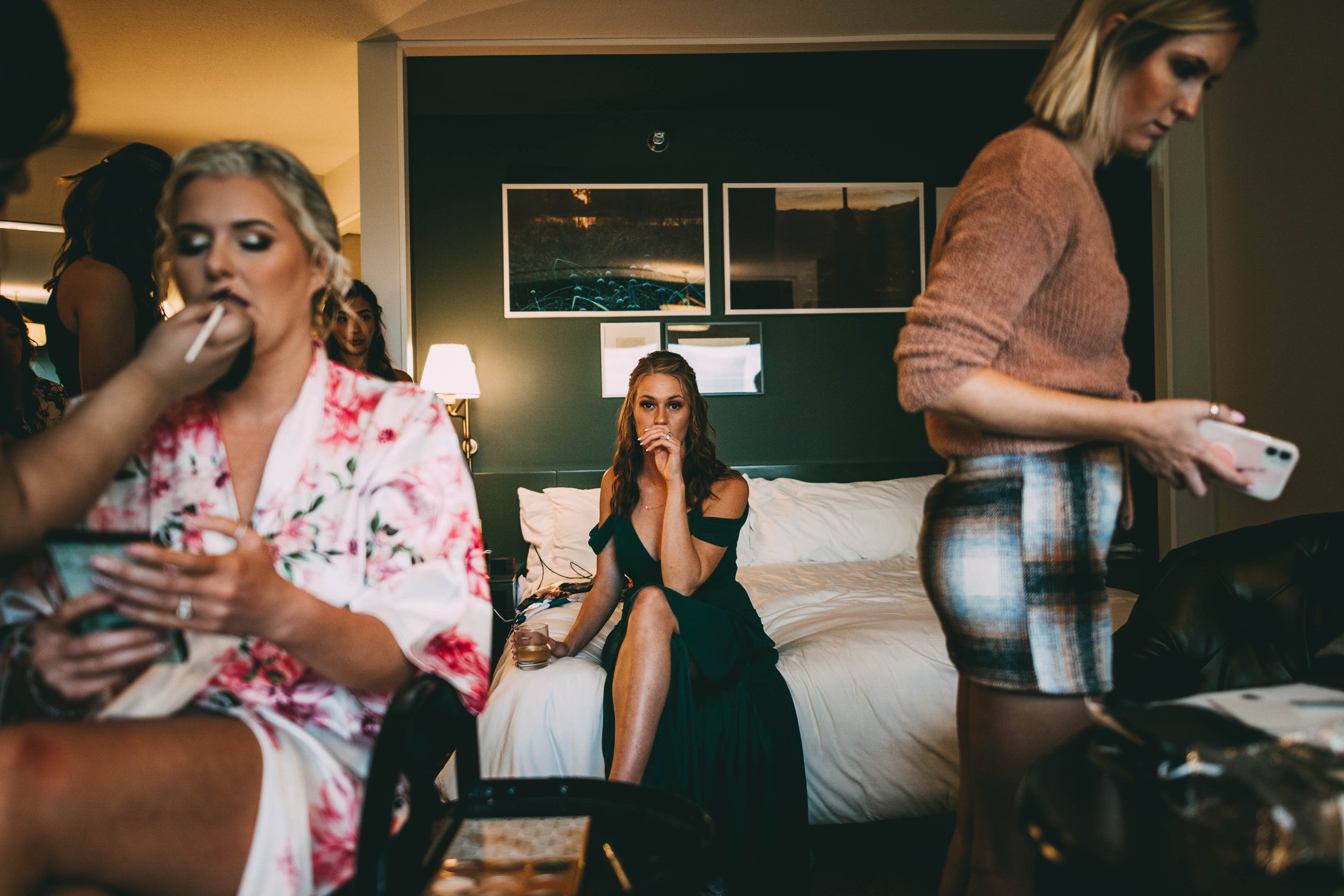 A woman in a black dress sitting on a bed with a drink in her hand, looking directly at the camera, while other women are around her, some using their phones, in a darkened hotel room with framed pictures on the wall.