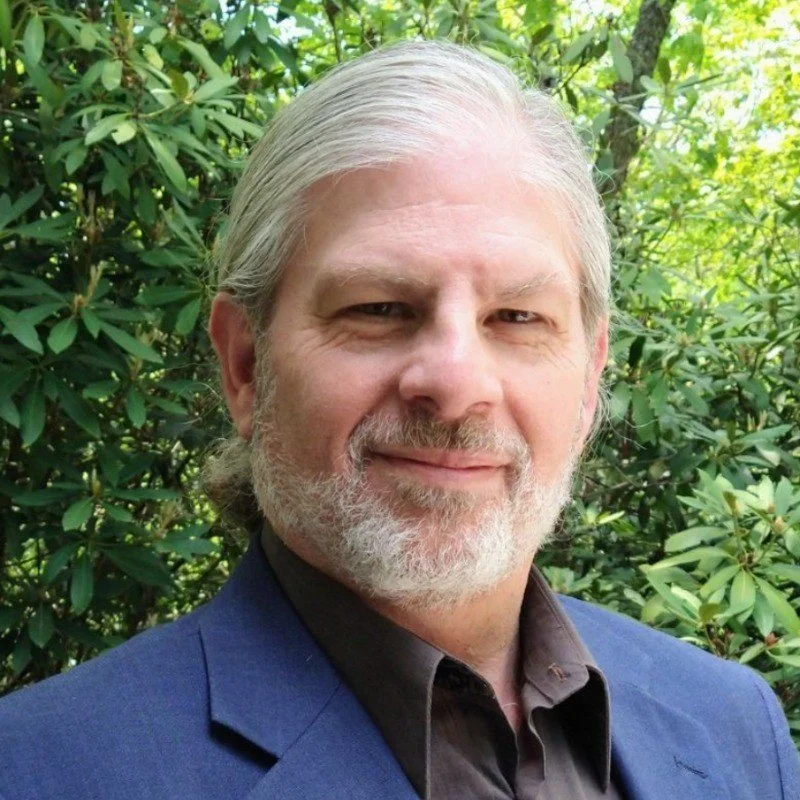 Portrait of a middle-aged man with gray hair and beard, smiling, wearing a dark dress shirt and a blue blazer, standing outdoors with green foliage in the background.