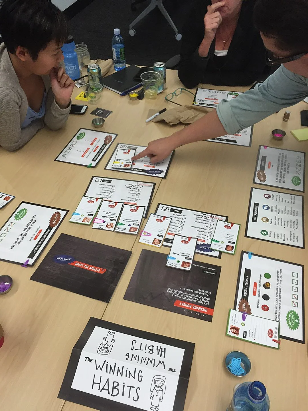 People playing a card game called 'Winning Habits' at a table filled with various game components, papers, and drinks.