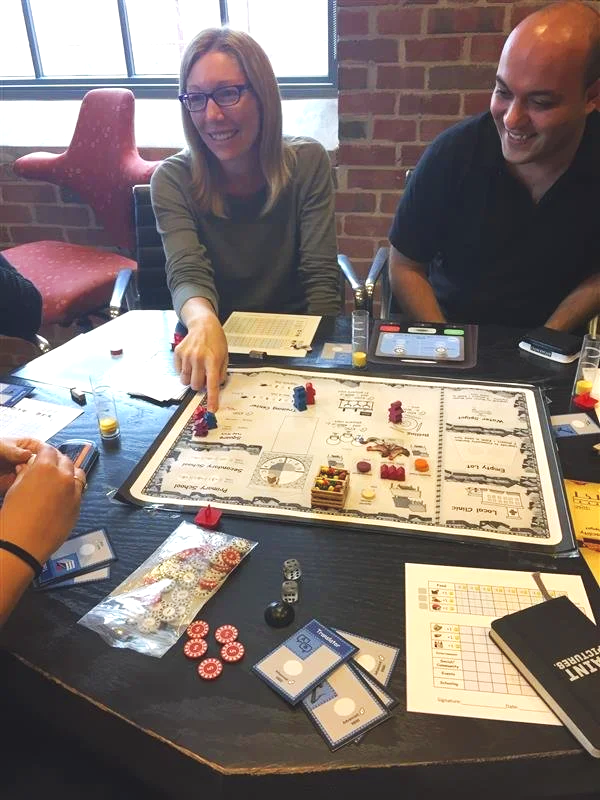 Two people playing Monopoly board game at a table with game pieces, cards, and dice, in a room with brick walls and a window.