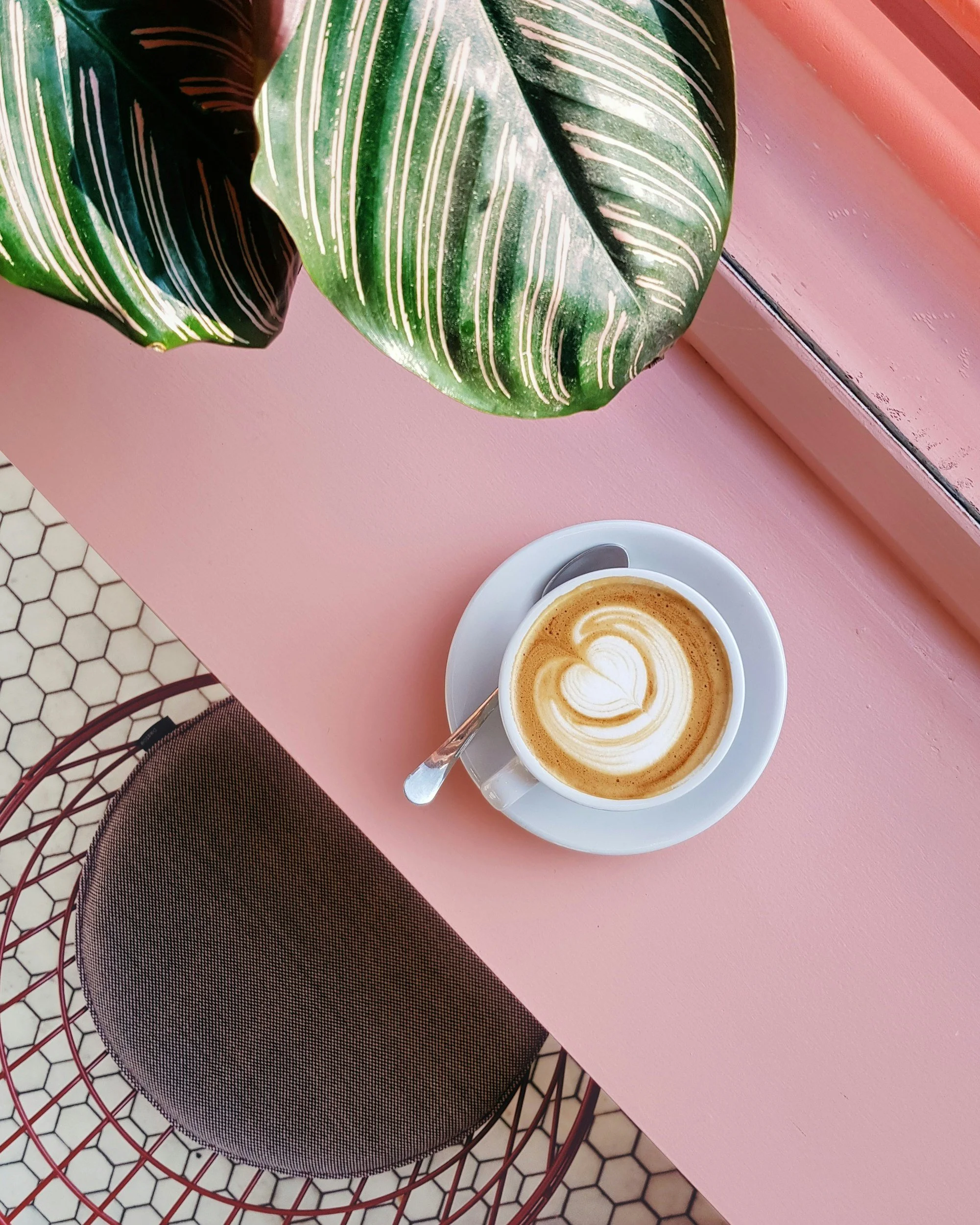 A cup of latte with heart-shaped foam art on a pink table next to a large green leaf plant, with a wire chair and hexagonal patterned floor visible.