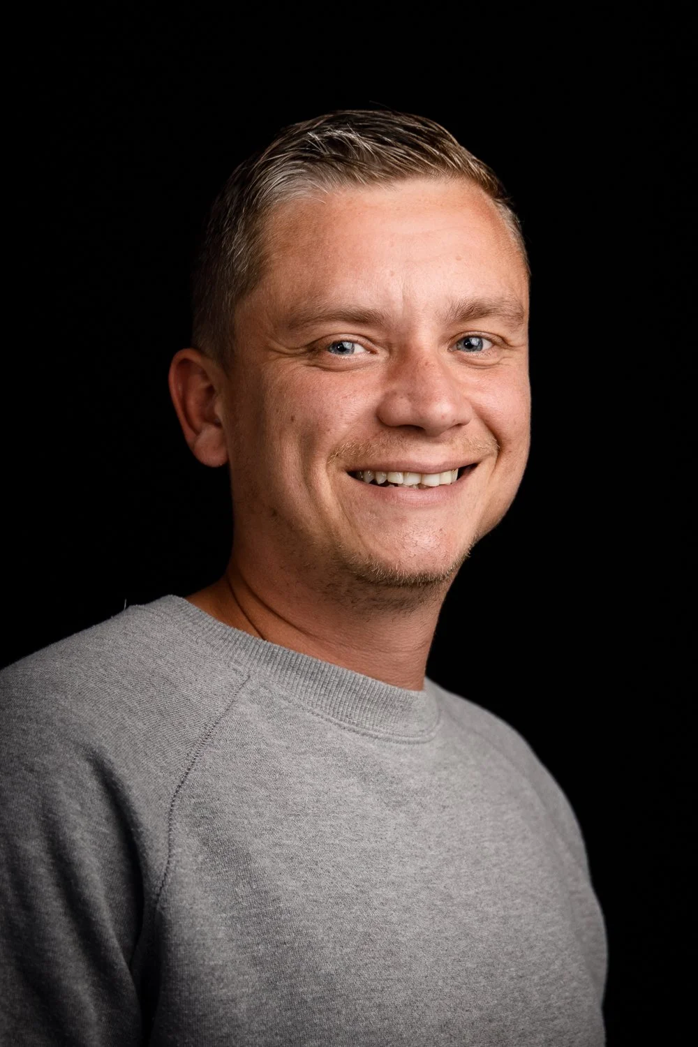 Smiling man with short hair and a gray sweater against a black background.