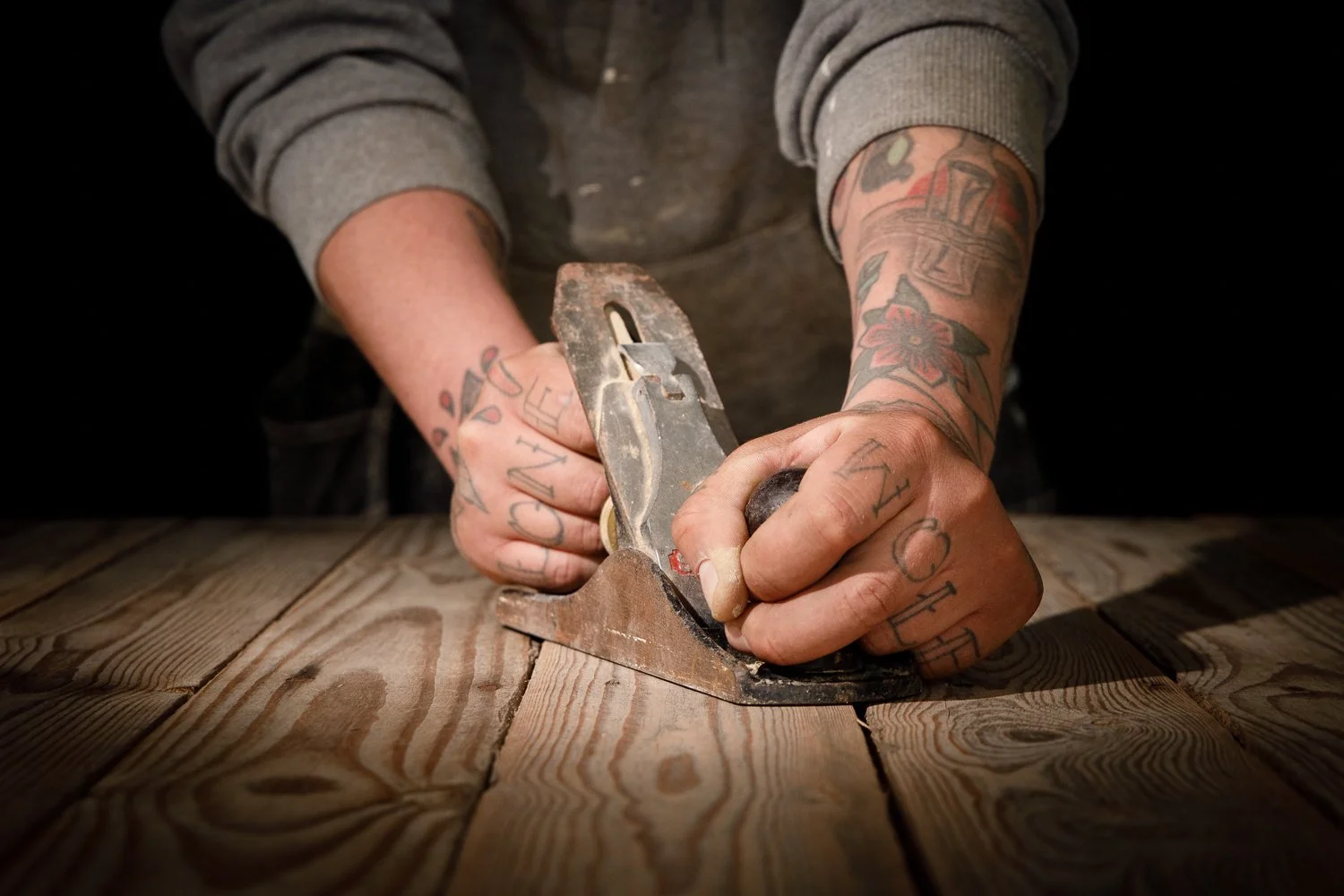 A person with tattooed arms using a hand planer to smooth a wooden surface.