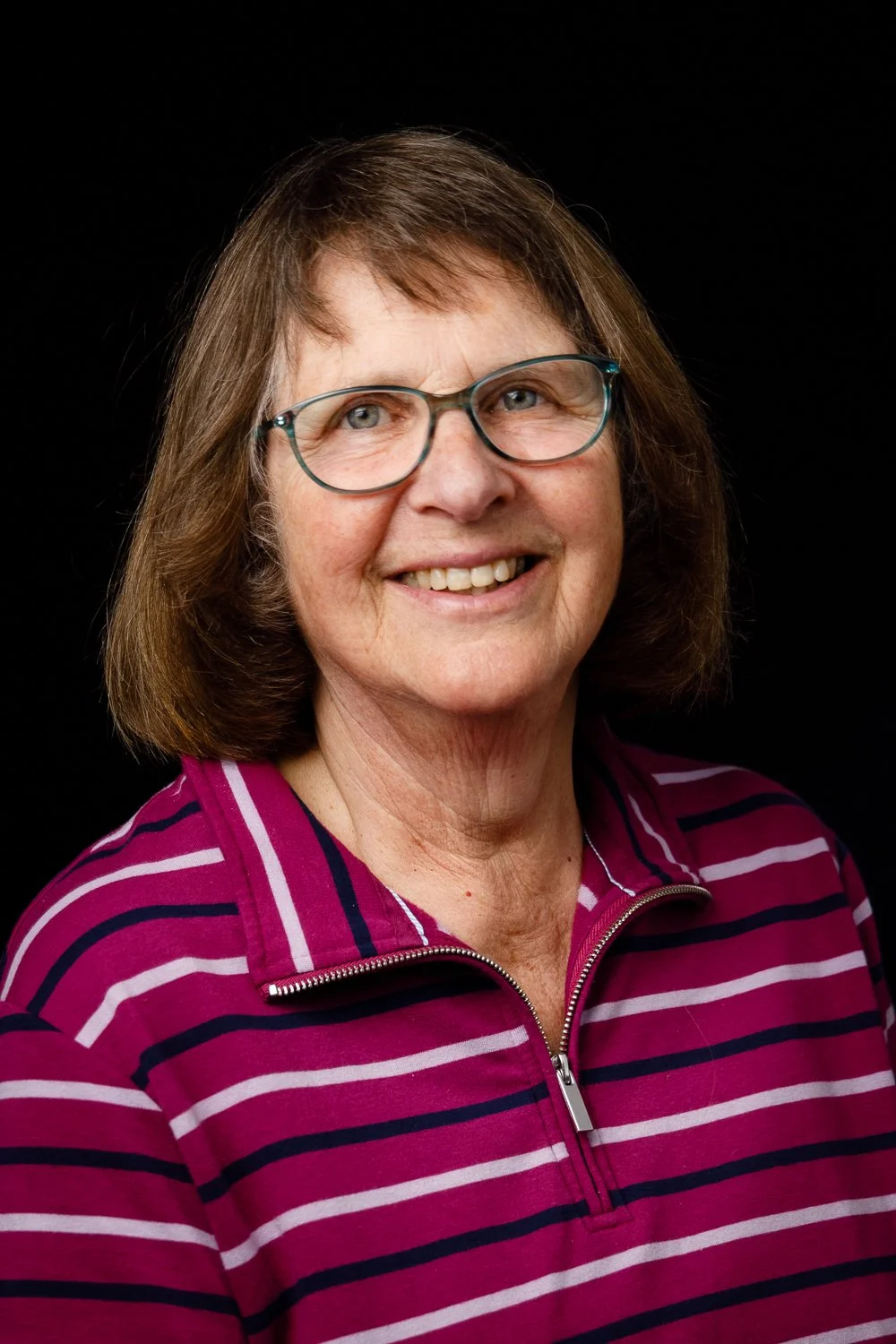 A middle-aged woman with shoulder-length brown hair, wearing glasses and a magenta and black striped shirt with a zip collar, smiling against a black background.