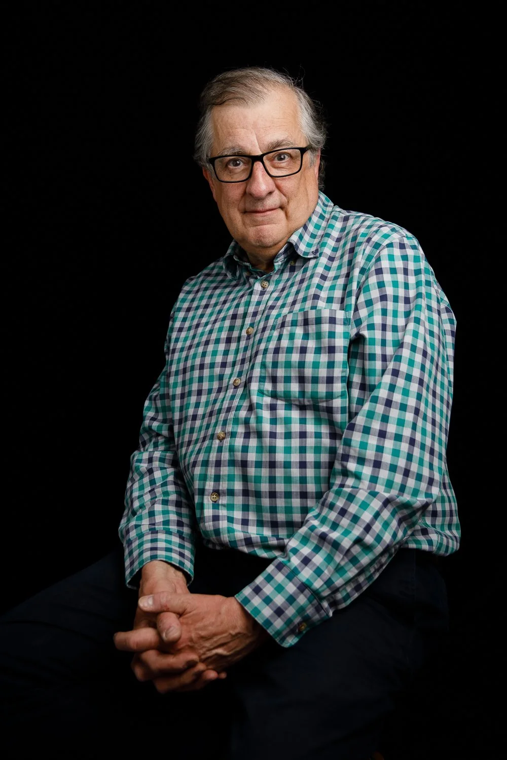 A mature man with glasses, wearing a green, blue, and white checkered shirt, sitting against a black background.