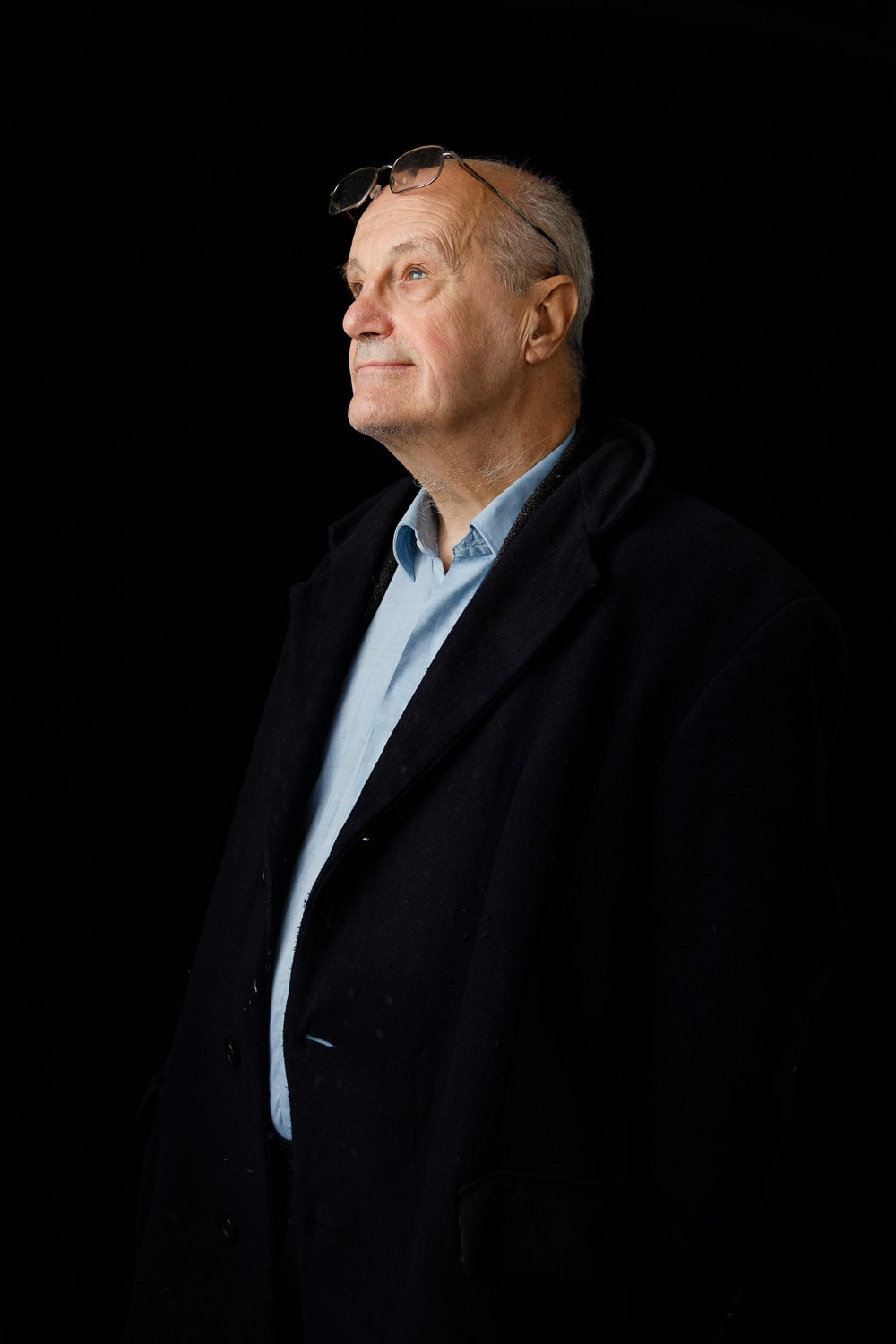 A senior man with glasses on his head, wearing a light blue shirt and dark coat, looking upwards against a black background.