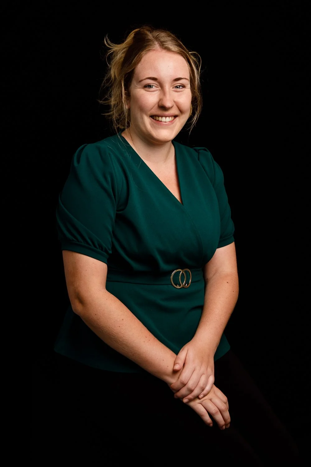A woman with blonde hair styled loosely, smiling while wearing a dark green V-neck dress with a decorative belt, posed against a black background.