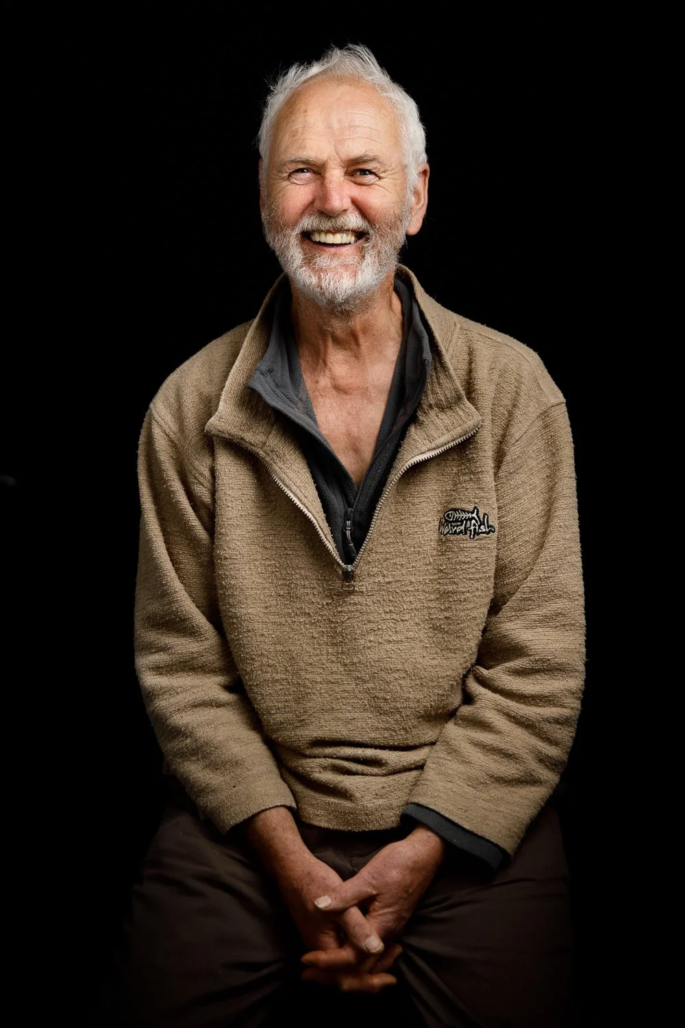 An elderly man with white hair and a beard, smiling, wearing a tan fleece jacket over a black shirt, sitting against a black background.