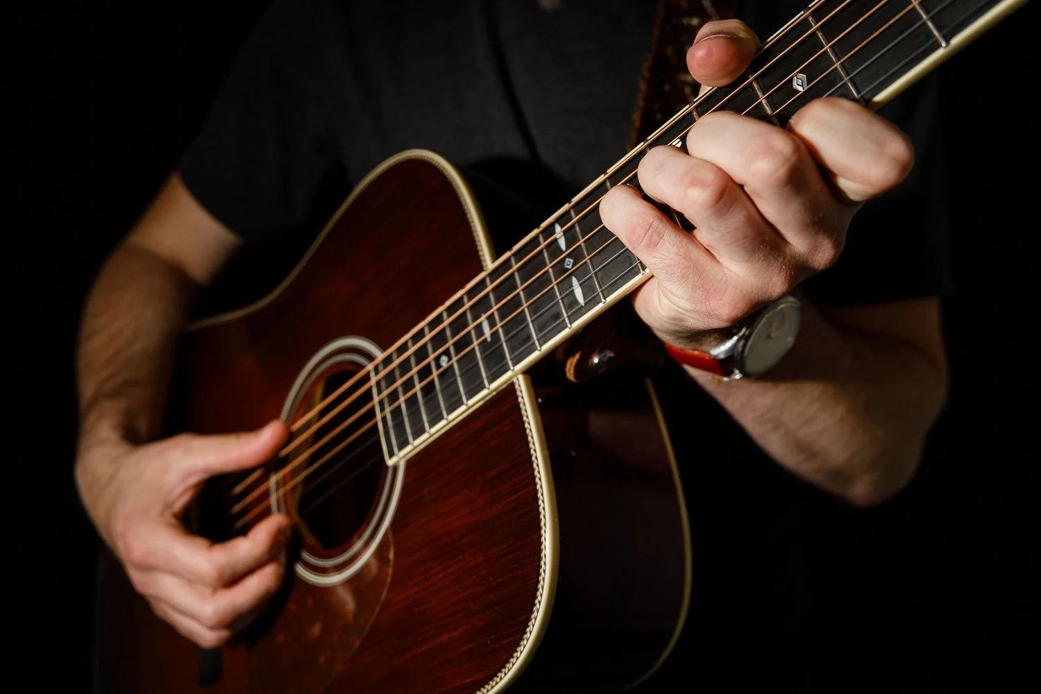 Close-up of a person playing an acoustic guitar, focusing on their hands and the guitar body against a dark background.