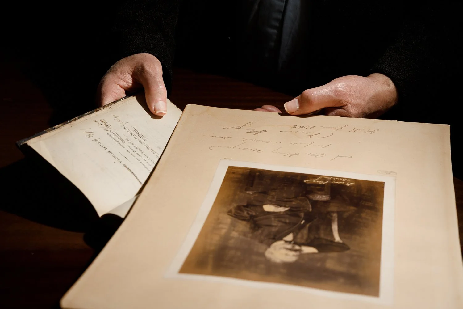 Person holding an open vintage album or scrapbook with a black-and-white photograph and handwritten notes on a dark wooden table.