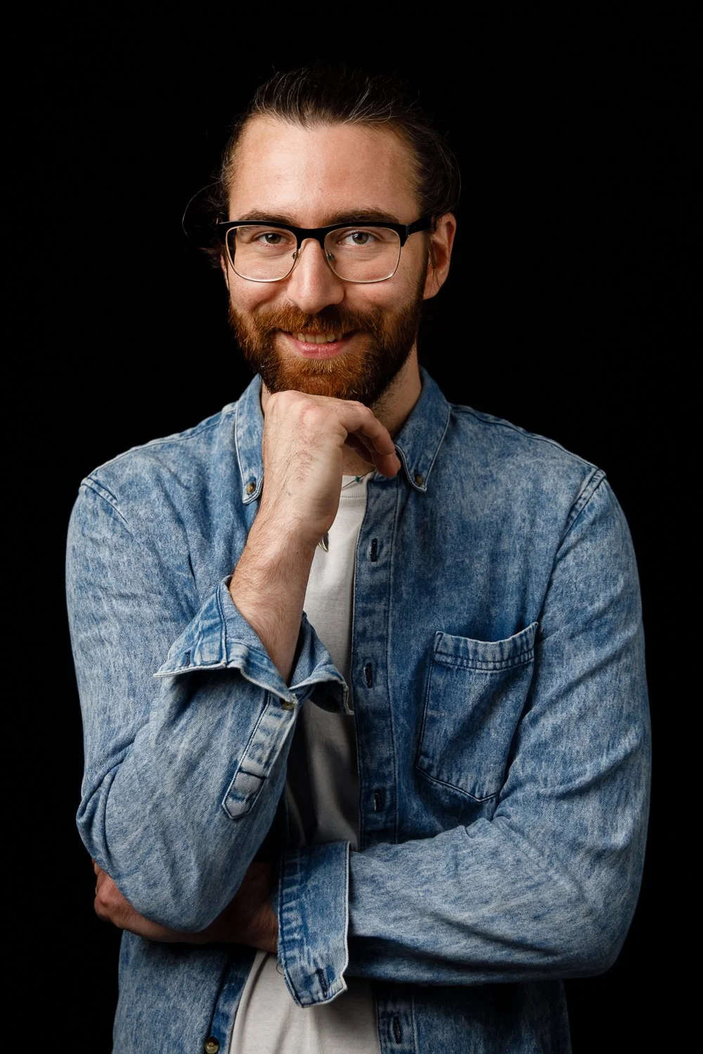 A young man with glasses and a beard smiling, wearing a denim shirt against a black background.