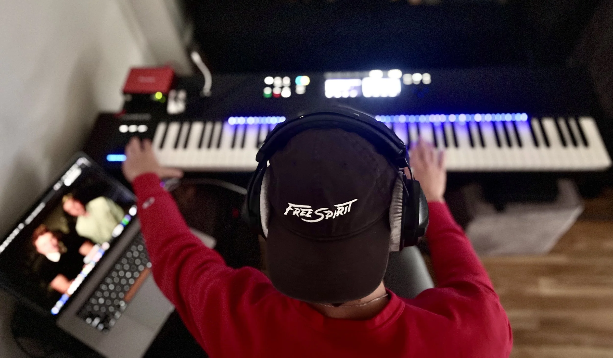 Top-down view of a person playing an electronic keyboard, wearing a black cap with 'Free Spirit' written on it and large headphones, with a laptop showing a video call nearby.