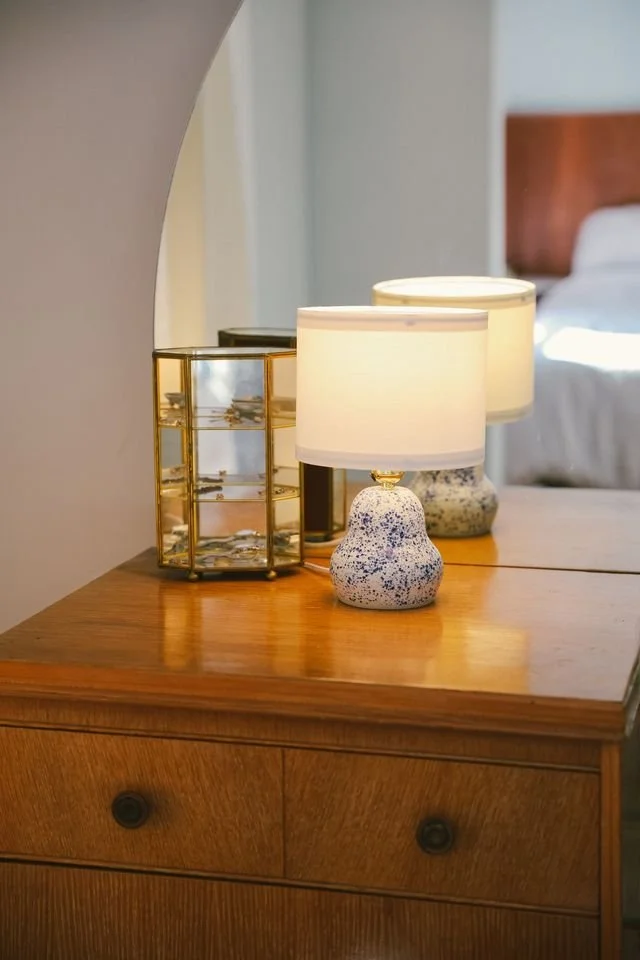 A table lamp with a speckled blue base and white shade, made by Natalya Jane on a wooden nightstand in a bedroom.