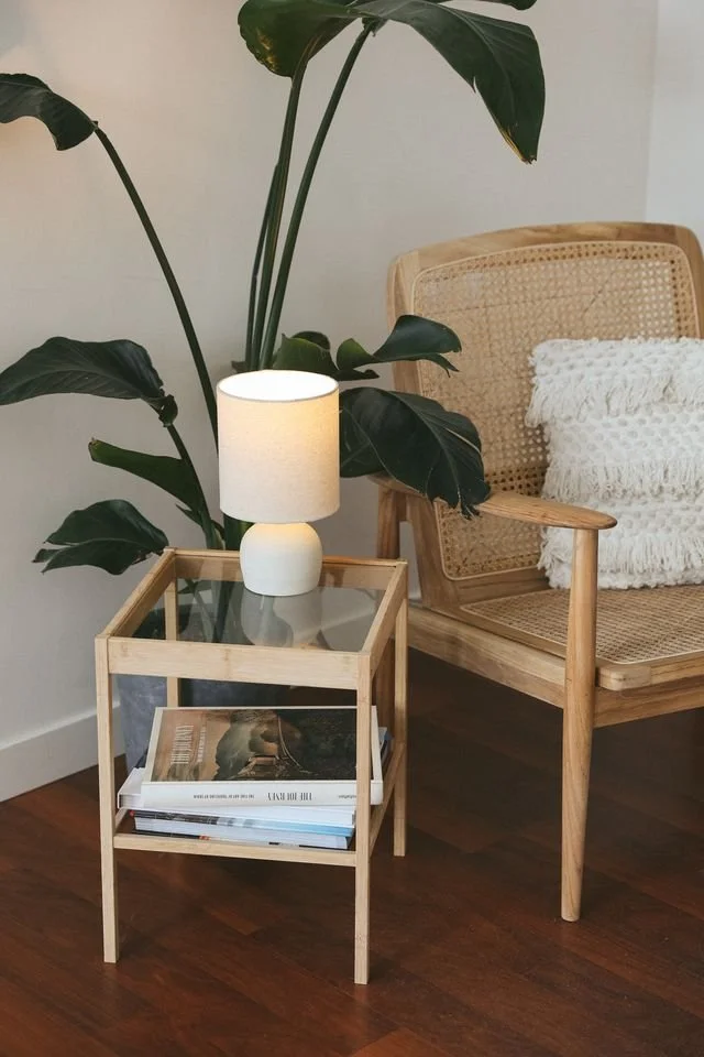 A living room corner with a wooden side table holding a white lamp made by Natalya Jane, a large green leafy plant behind it, and a wooden chair with a woven cane backrest and a white textured throw pillow.