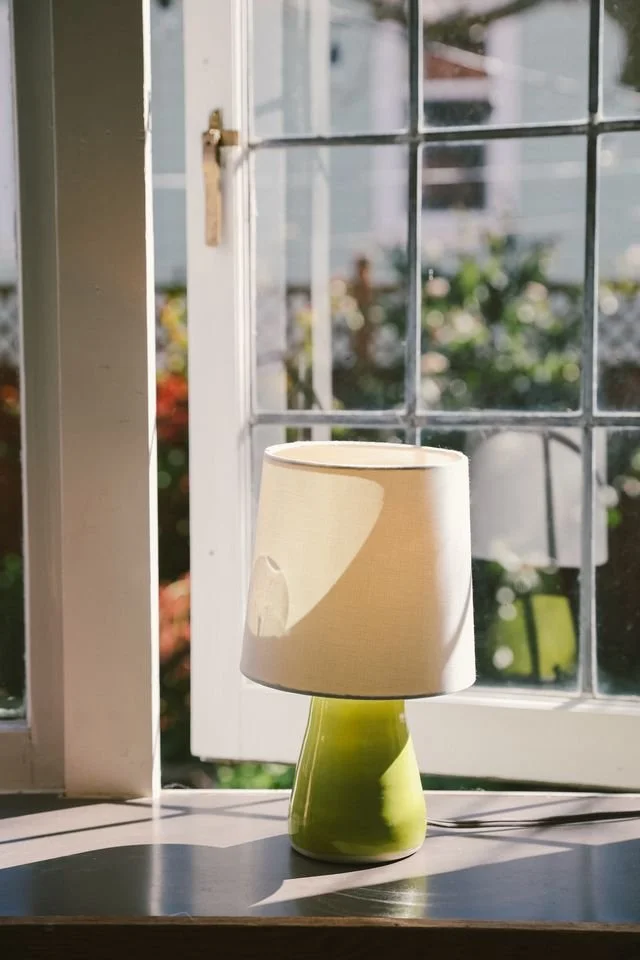 A green ceramic table lamp with a beige lampshade made by Natalya Jane sitting on a windowsill, with sunlight casting shadows.