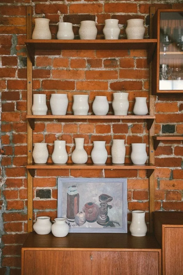 Interior wall with brick background and wooden shelves displaying white ceramic vases made by Natalya Jane and a framed painting of pottery on a wooden cabinet.