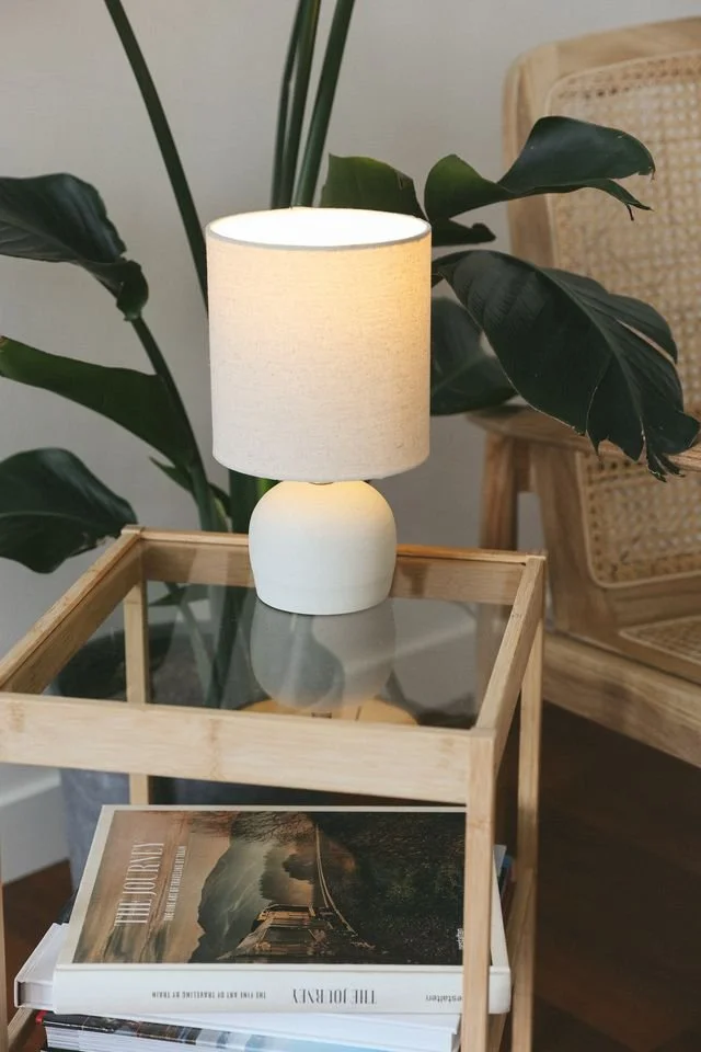 A white table lamp with a round base and shade, made by Natalya Jane, sitting on a wooden and glass side table. Large green leafy plants are in the background, with a rattan chair nearby.