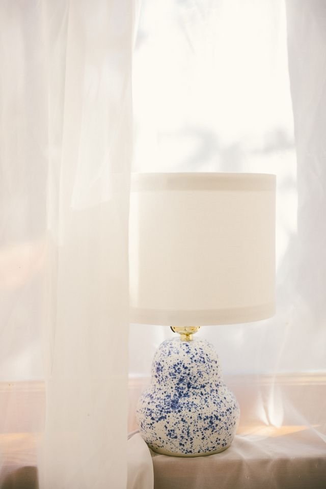 A table lamp with a speckled blue and white ceramic base and a white lampshade made by Natalya Jane, placed on a table near sheer white curtains.