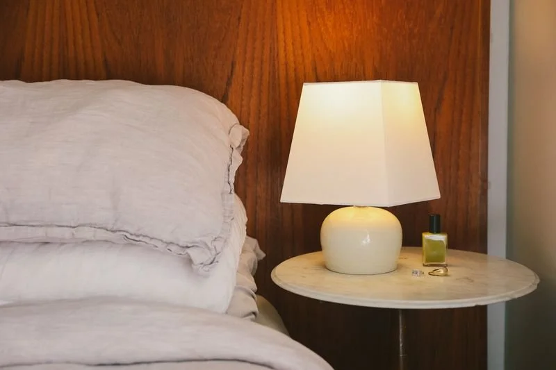 A bedroom nightstand with a lit table lamp made by Natalya Jane, and a small bottle of nail polish sit next to a bed with white linens and a wooden headboard.