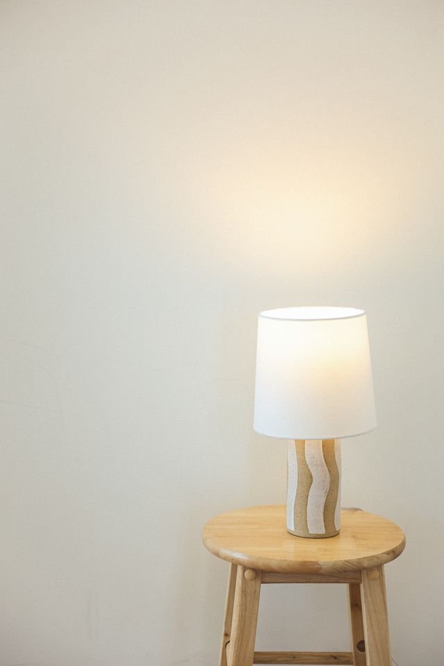 A table lamp with a white shade and a wavy, textured beige base, made by Natalya Jane, on a wooden stool against a plain, light-colored wall.